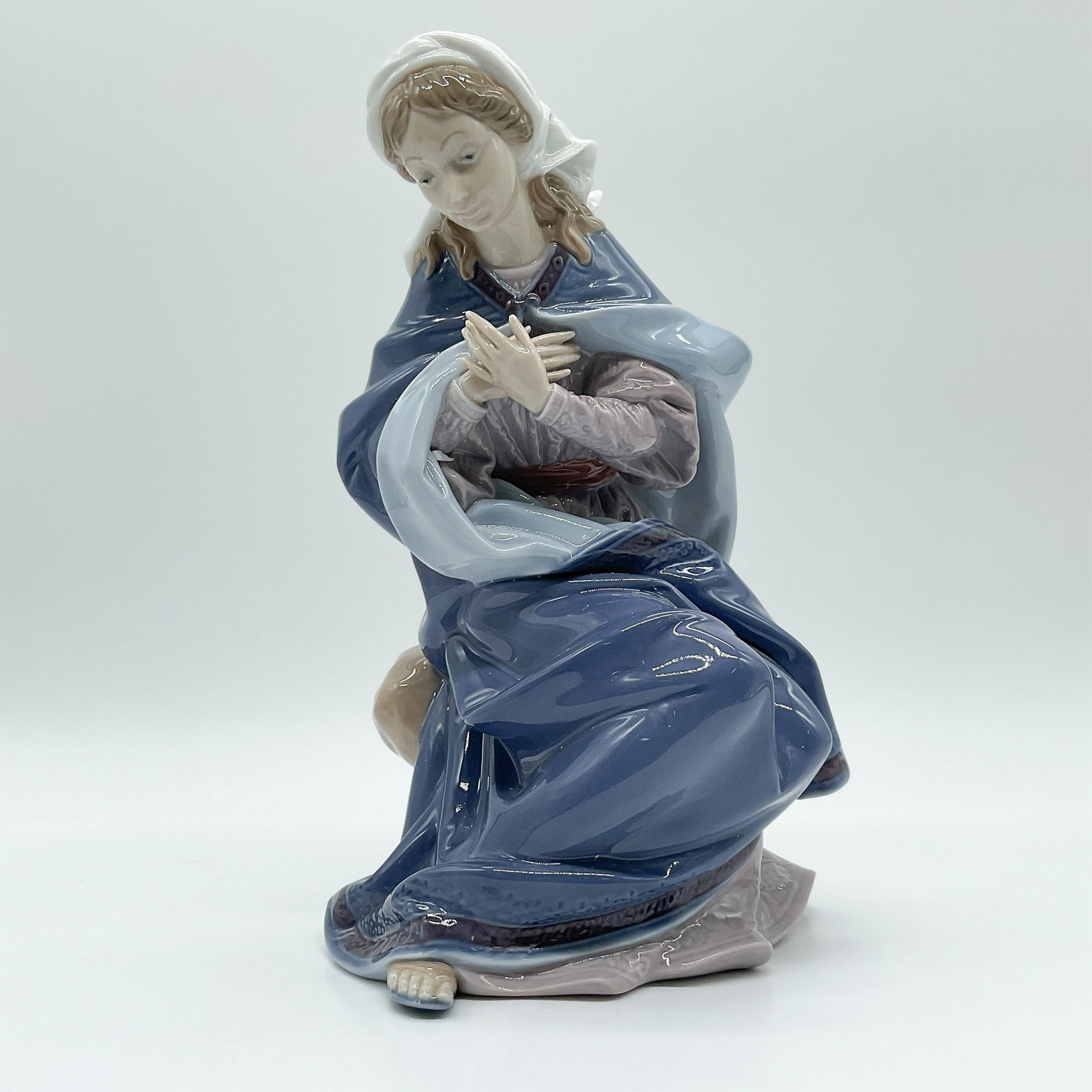 Lladro Porcelain Figurine, Virgin Mary 1001387: The figurine depicts the Virgin Mary in a serene seated pose, adorned with a flowing blue mantle and delicate white veil. The soft, muted tones and elegant design highlight the skill of Lladro's artis