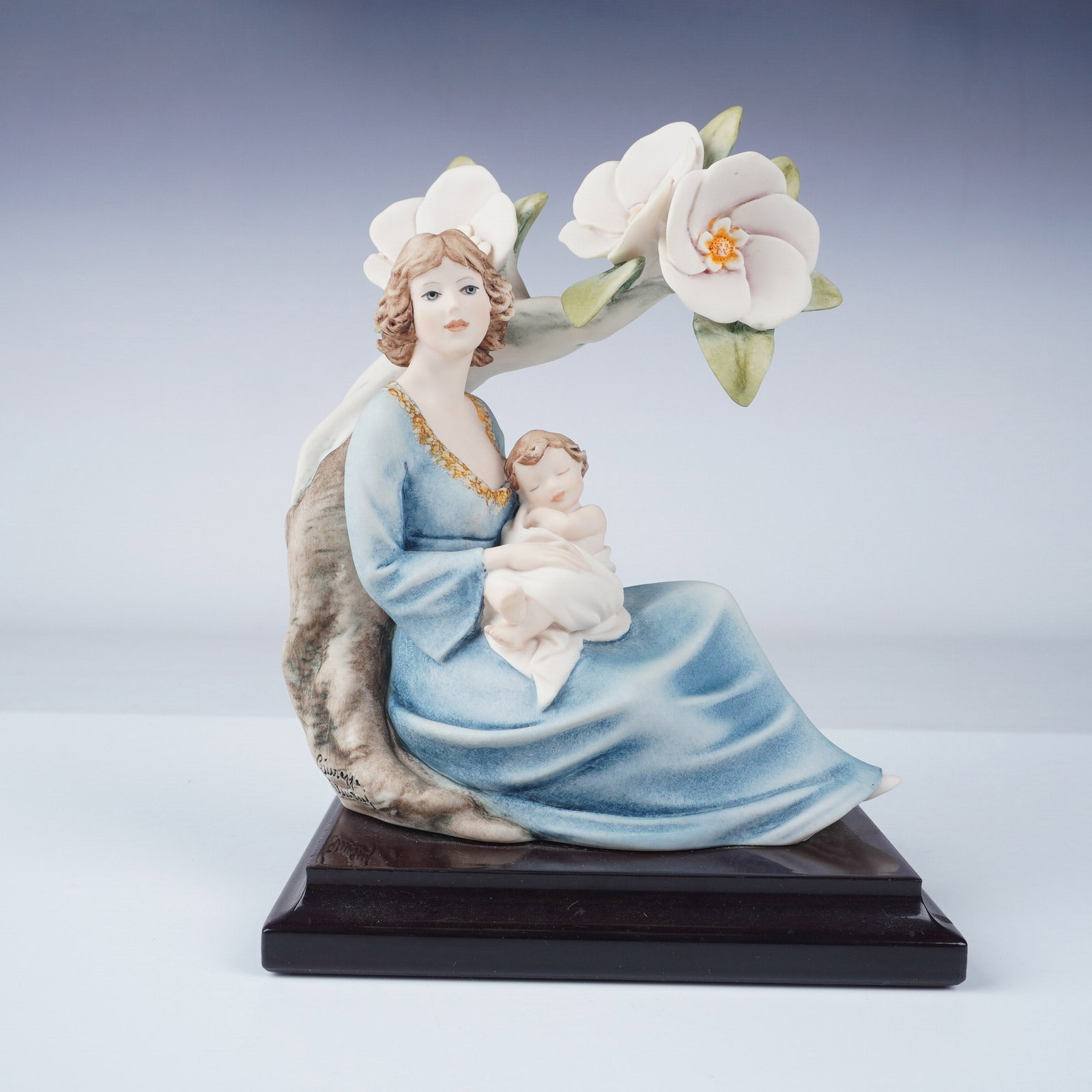 Florence Giuseppe Armani Blossoms Porcelain Figurine: A finely crafted Florence porcelain figurine by Giuseppe Armani, titled Blossoms, depicting a serene mother in a flowing blue gown cradling her infant under a flowering magnolia tree. This piece showc