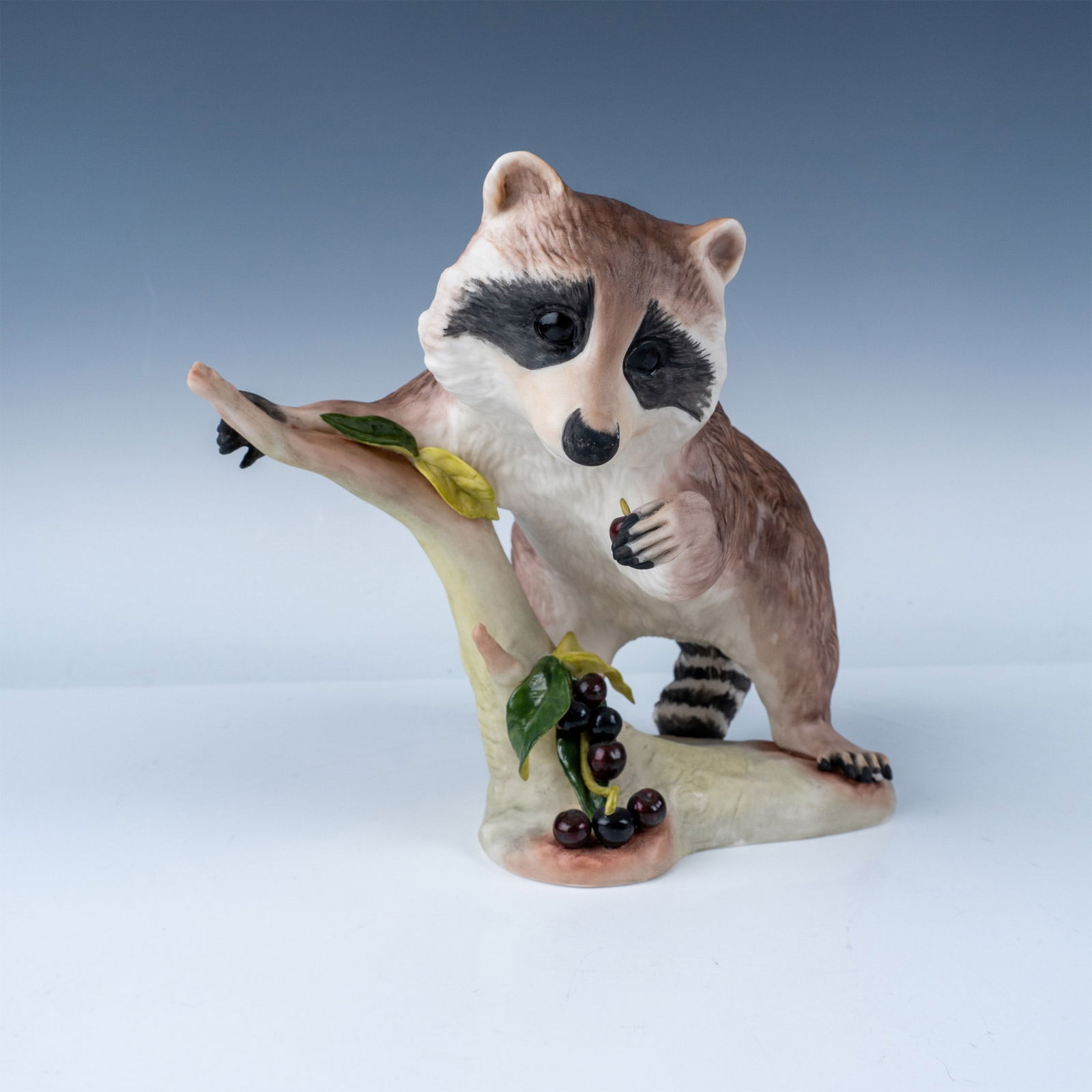 Cybis Porcelain Figurine: "Raffles" the Raccoon: Presenting "Raffles" the Raccoon, a charming porcelain figurine crafted by the renowned Cybis studio. Introduced in 1965, this piece showcases a raccoon perched on a tree branch, indulging in a cluste