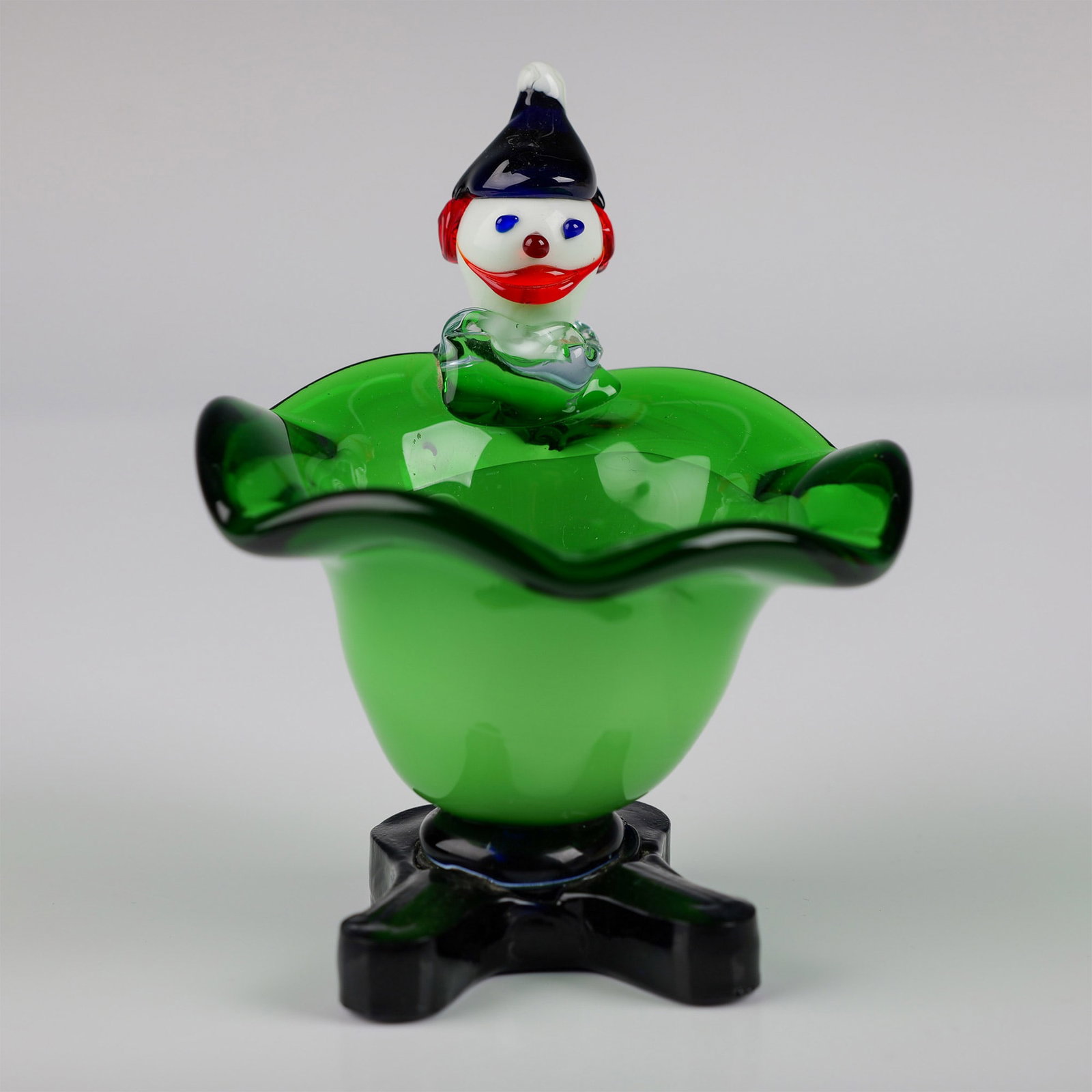 Murano Glass Green Bowl with Figural Clown Accent: A charming Murano glass decorative bowl featuring a vibrant green design with an elegantly ruffled edge. The centerpiece is a whimsical clown figure with colorful details, adding a playful touch. The 