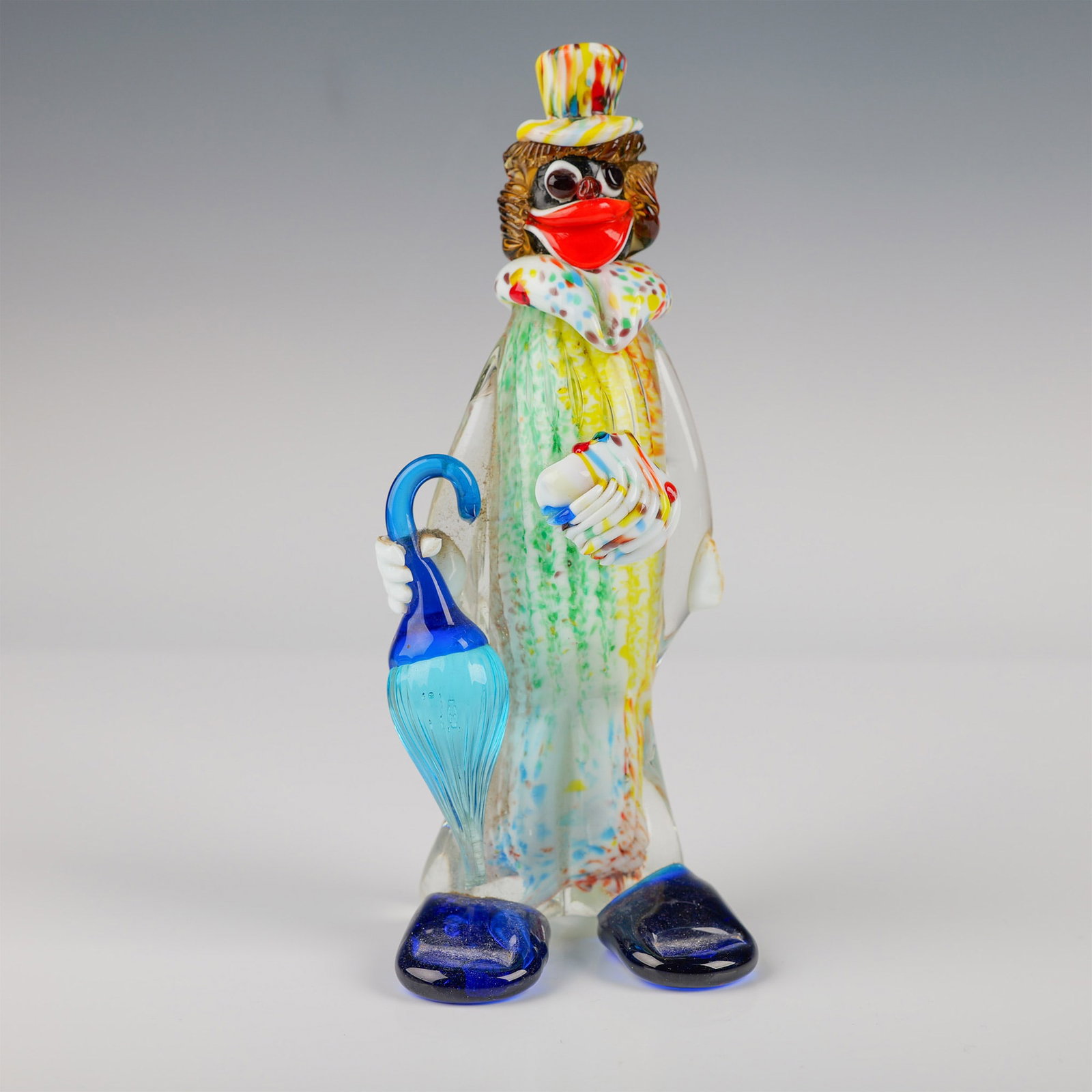 Murano Glass Clown Figurine with Umbrella: This charming Murano glass clown figurine stands at 8.75" tall, featuring a playful design with a colorful array of hues. The clown is adorned with a multi-colored top hat, a ruffled collar, and holds
