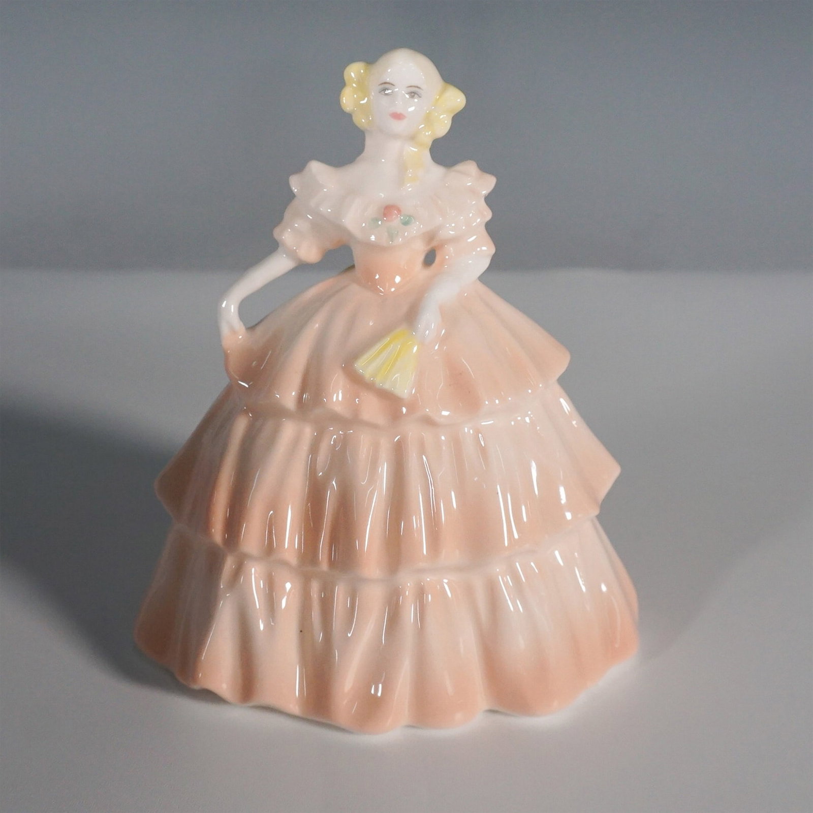 Coalport Minuettes Summertime Figurine: This Coalport Minuettes Summertime figurine features a charming lady in a peach-colored, tiered gown, delicately holding a yellow fan. Hand-decorated and modelled by Martin Evans, this figurine reflec