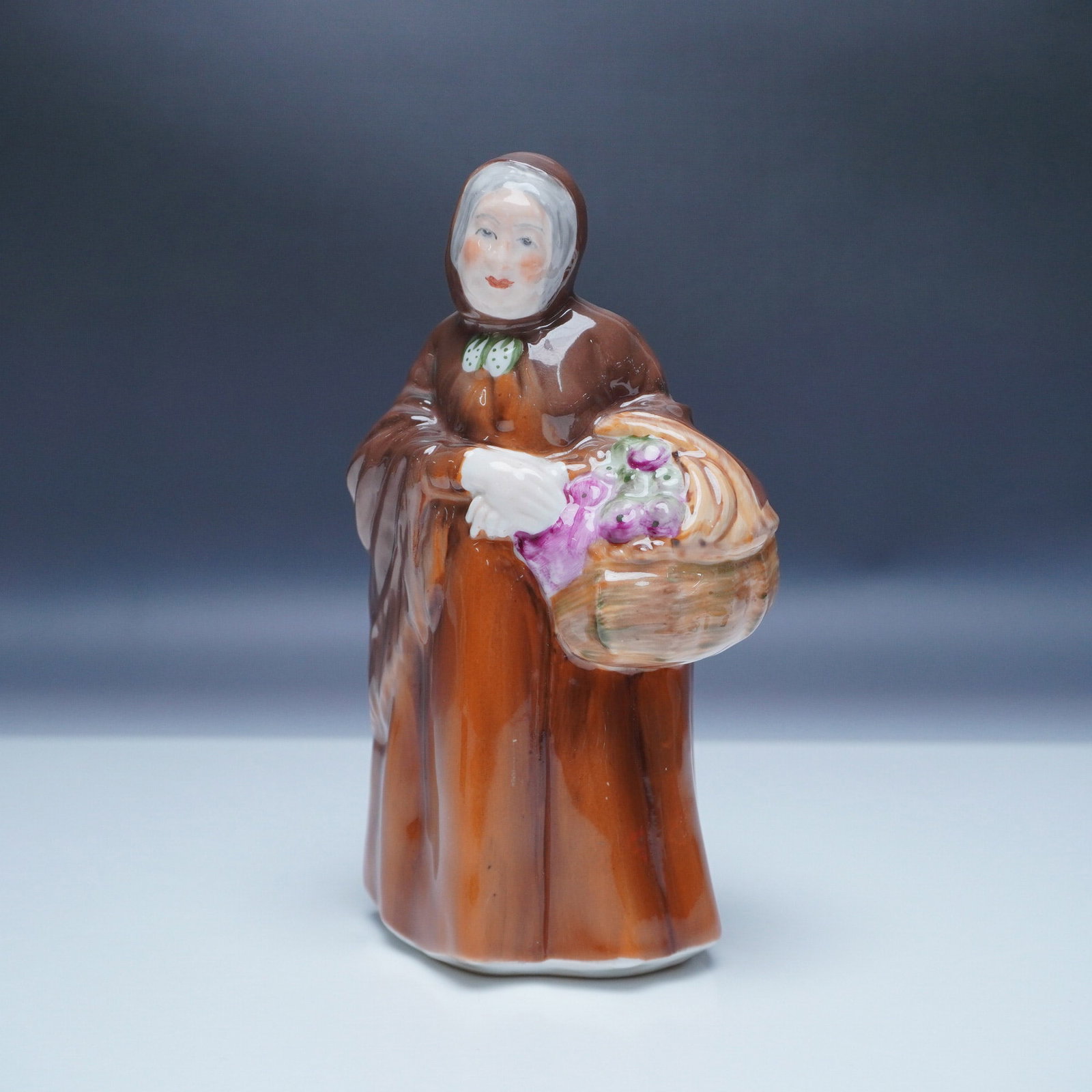 Coalport Figurine Old Market Lady: This charming Coalport figurine, "Old Market Lady," showcases the timeless elegance of fine English porcelain craftsmanship. Standing at 6.5"H, this exquisite piece depicts an elderly woman in a brown