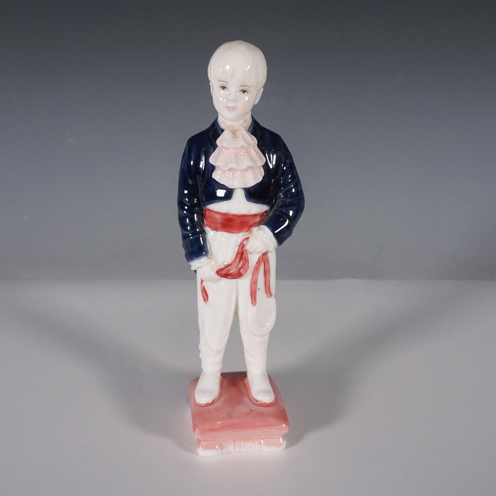 Coalport Porcelain Figurine, Page Boy: This Coalport Page Boy figurine captures the charm of a young boy dressed in a formal page boy outfit, featuring a navy blue jacket, ruffled pink shirt, white trousers, and a red sash. Made from fine 