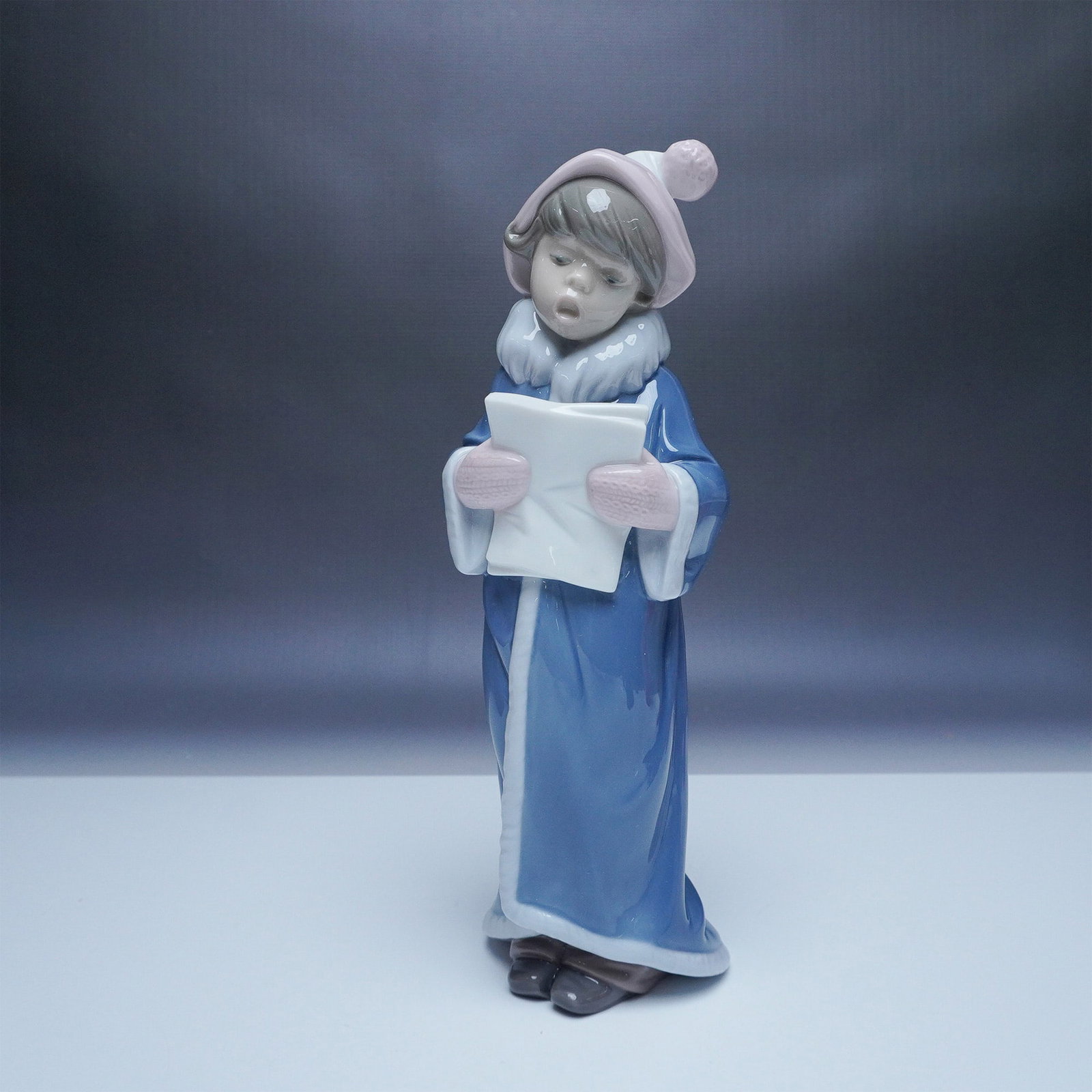 Lladro Christmas Caroler Figurine 01006533: Celebrate the holiday season with this charming Lladro porcelain figurine titled Christmas Caroler, model number 01006533. The figurine features a young boy dressed warmly in a blue winter coat with w