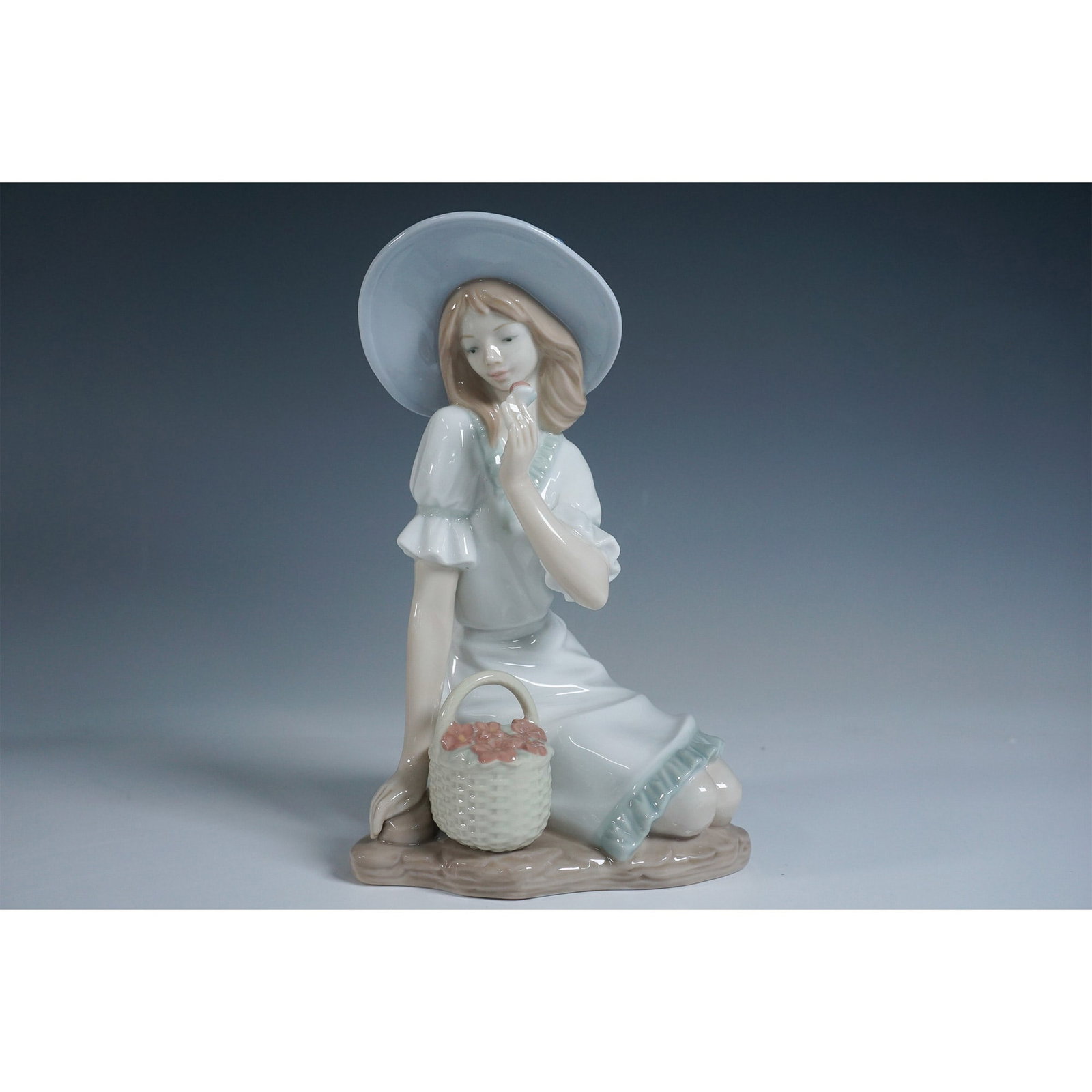 Nao by Lladro Porcelain Figurine, Girl with Basket 1305: A charming Nao by Lladro porcelain figurine titled "Girl with Basket," model number 1365. This delicate piece depicts a young girl in a flowing white dress with pastel accents, wearing a wide-brimmed 