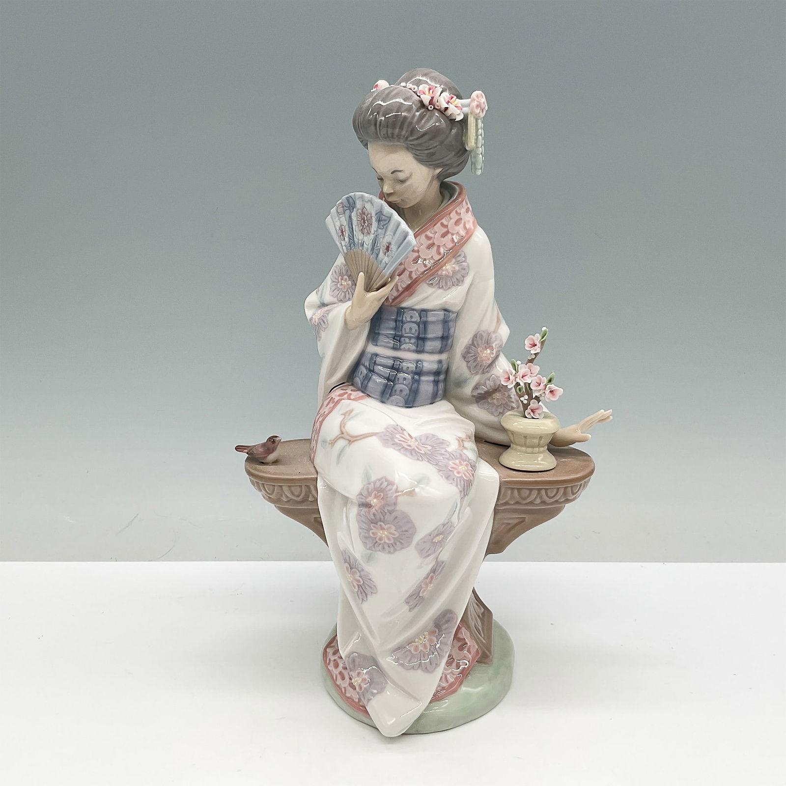 Nippon Lady 01005327 - Lladro Figurine: Elegant Lladro porcelain figurine titled Nippon Lady (01005327), depicting a seated Japanese woman dressed in a beautifully adorned kimono, gracefully holding a folding fan while seated on an ornate b