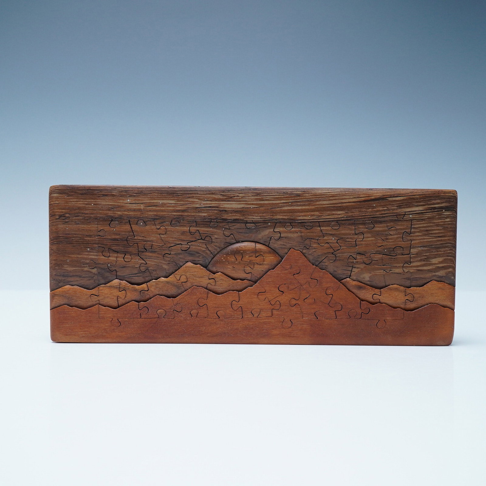 Handcrafted Wooden Puzzle Art, Mountain Landscape: A beautifully handcrafted wooden puzzle featuring an intricately designed mountain landscape with a rising or setting sun. The puzzle is seamlessly embedded into a rectangular wooden frame, creating a