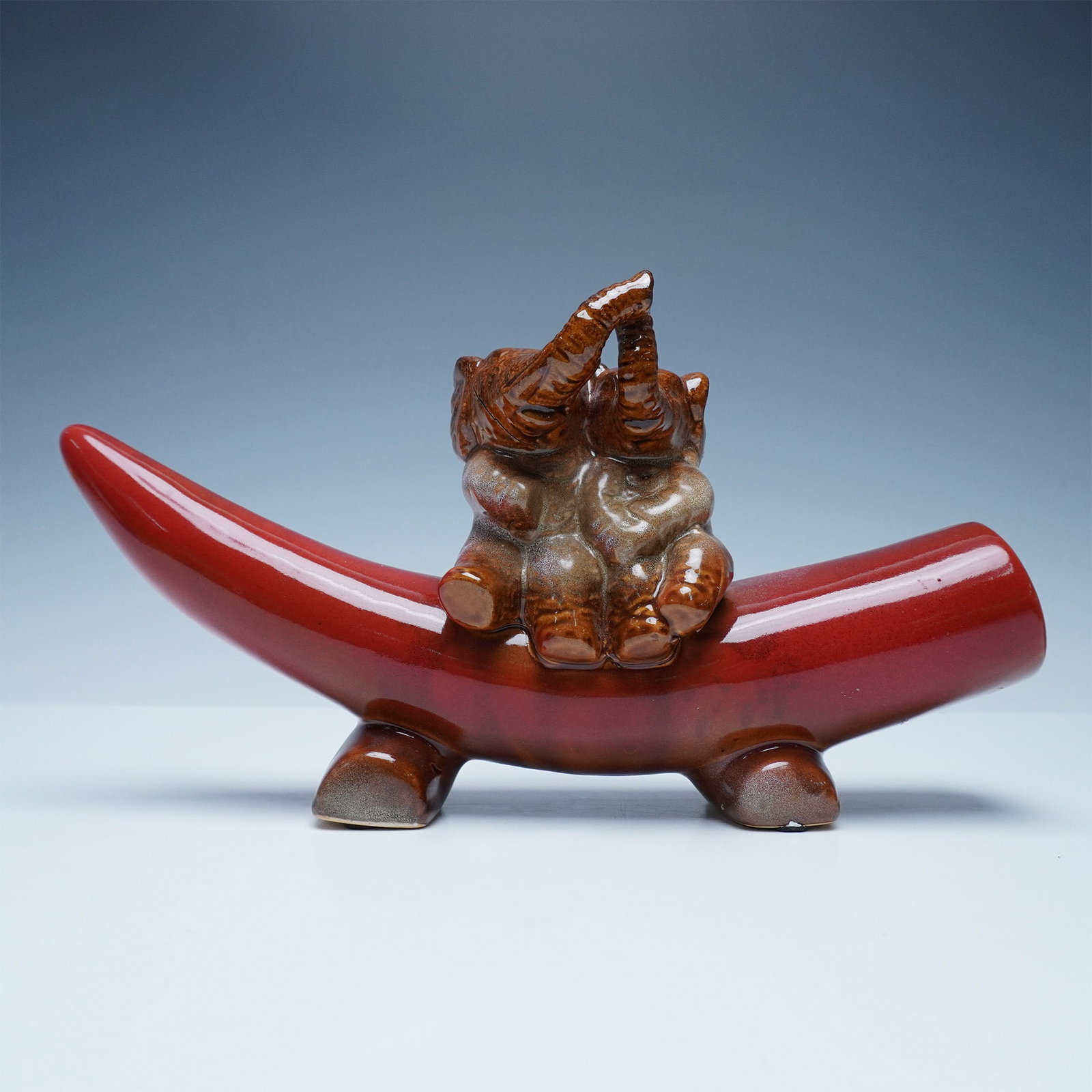 Glazed Ceramic Elephant Figurine on Horn Styled Base: A charming glazed ceramic figurine featuring two playful elephants sitting atop a horn styled base. The elephants, depicted in a warm brown glaze with detailed sculpting, raise their trunks in an expr