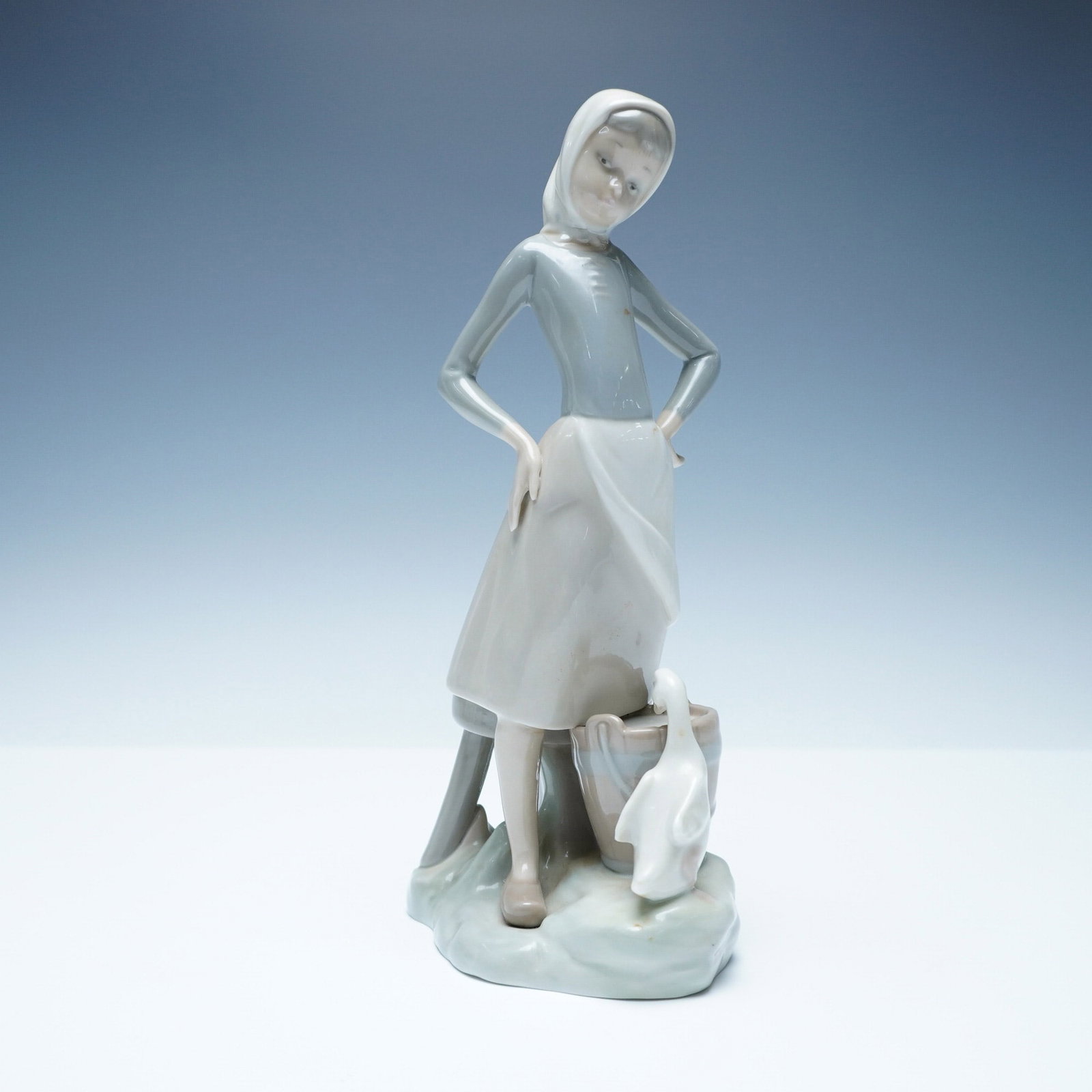 Lladro Figurine, Girl with Milk Pail 1004682: This Lladro figurine, titled Girl with Milk Pail (1004682), depicts a young girl in a bonnet and apron standing beside a wooden pail with a curious goose peering in. Sculpted in Lladro?s signature sof