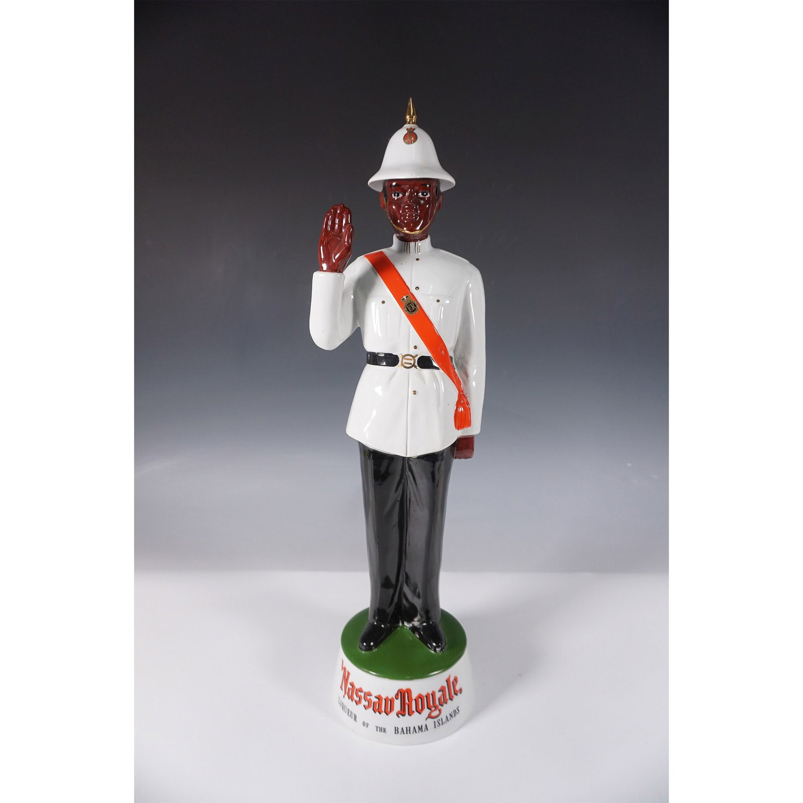 Nassau Royale Ceramic Liqueur Decanter, Bahamian Policeman: This Nassau Royale ceramic liqueur decanter depicts a Bahamian policeman in full ceremonial uniform, complete with a removable white pith helmet featuring a gold finial. The figure is dressed in a whi