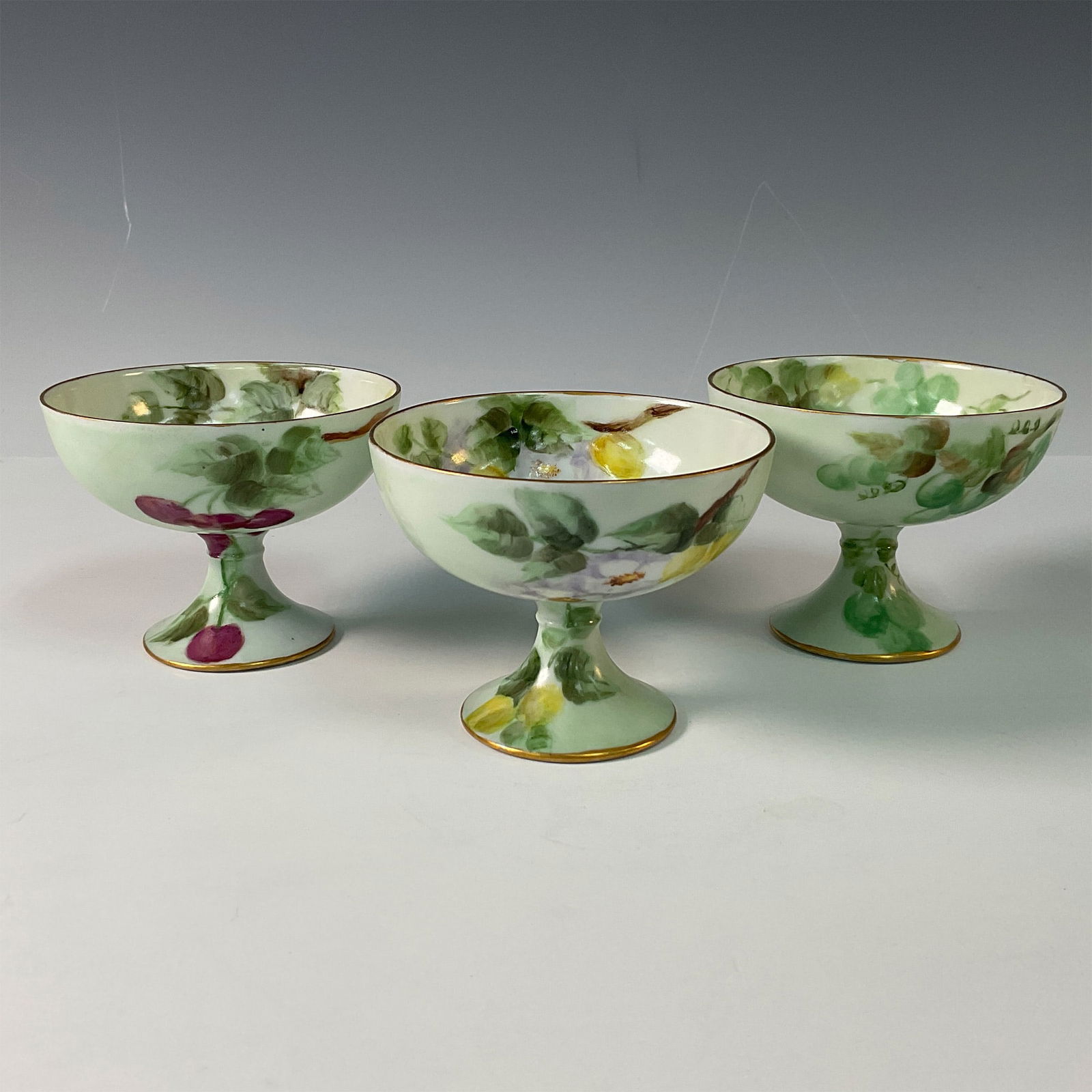 3pc Oscar & Edgar Gutherz Fruit Motif Punch Cups: A charming set of 3 Oscar & Edgar Gutherz porcelain punch cups, each hand painted with unique fruit motifs. The designs include vibrant depictions of grapes, cherries, and lemons, accented with detail
