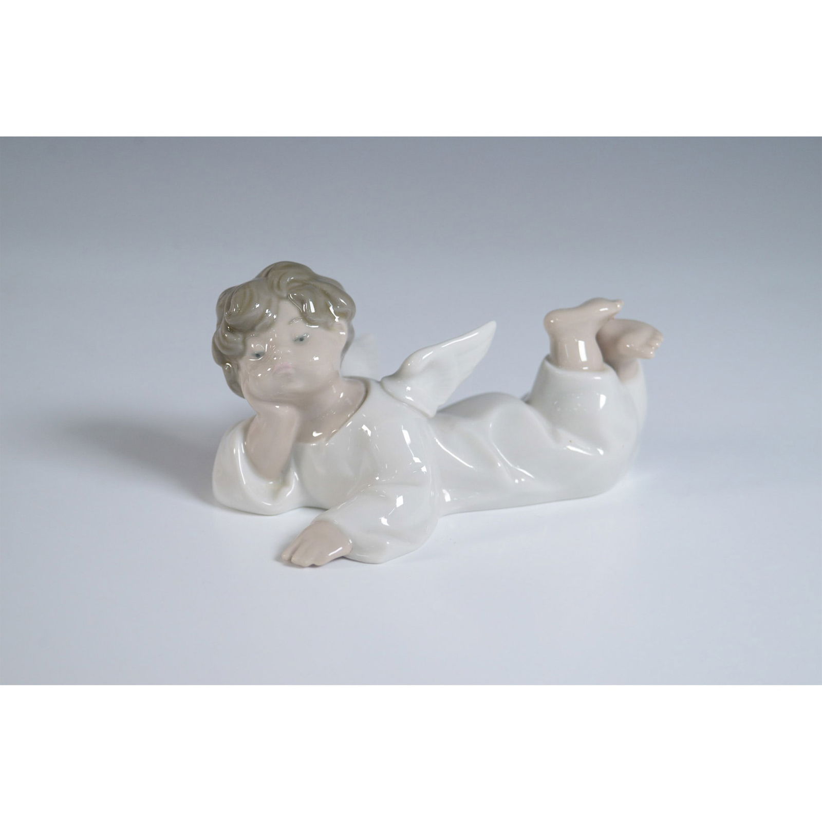 Lladro Porcelain Figurine, Angel Reclining 1004541: Lladro porcelain figurine, titled Angel Reclining (1004541), depicts a peaceful angel lying serenely, gazing gently with delicate features. Sculpted by Fulgencio Garcia, the figurine's smooth, flowing