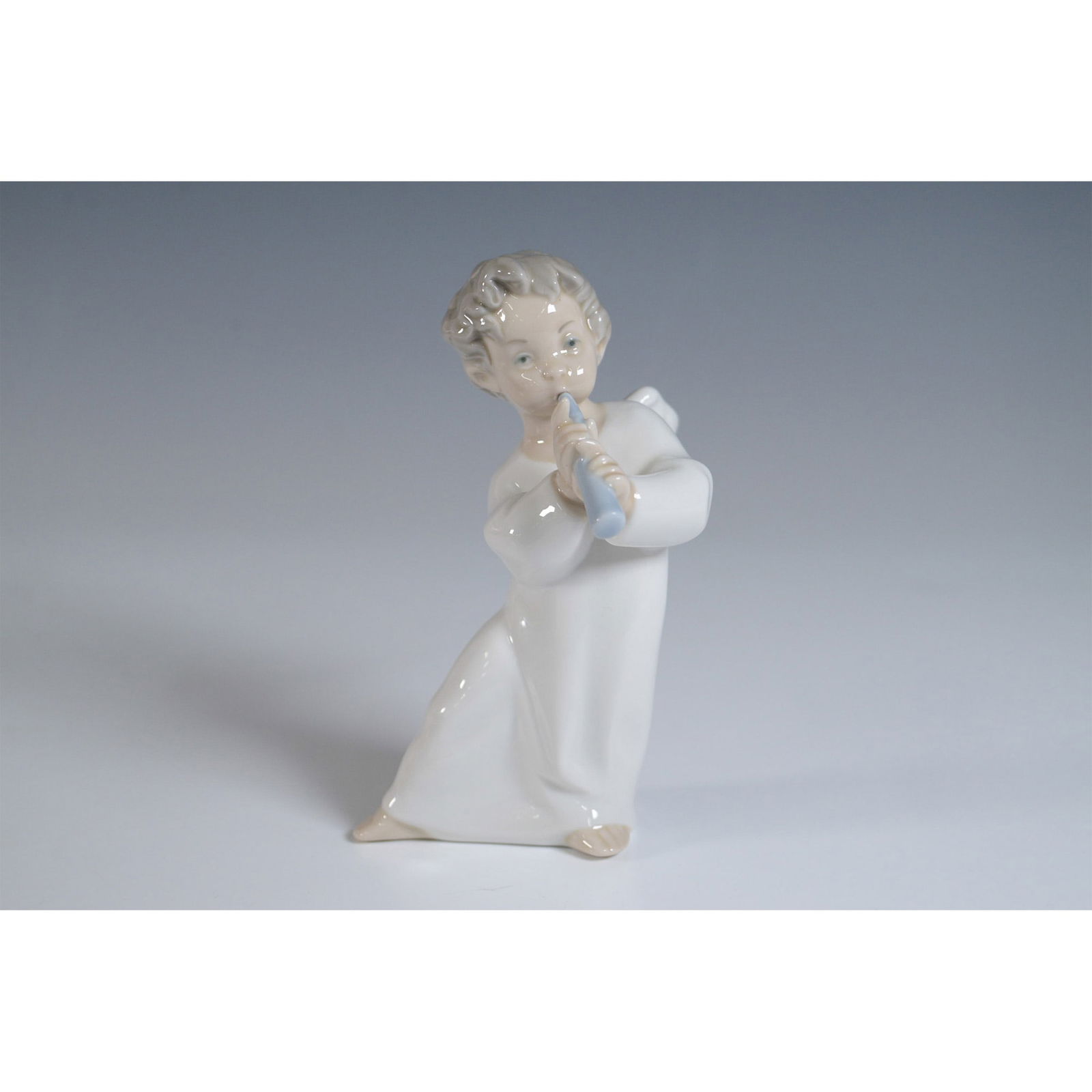 Lladro Porcelain Figurine, Angel with Flute 1004540: A charming Lladro porcelain figurine titled Angel with Flute, model number 1004540. This delicate piece depicts a young angel in a flowing white gown, playing a pastel blue flute. The figurine showcas