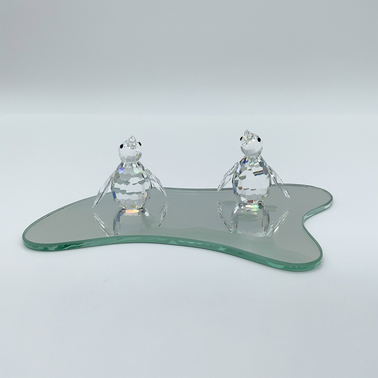 Swarovski Crystal Figurine Penguin Pair with Mirror Base: This charming Swarovski Crystal Penguin pair, part of the "Kingdom of Ice and Snow" theme, features clear crystal penguins with black eyes, mounted on a sleek mirror base. Designed by Max Schreck, thi