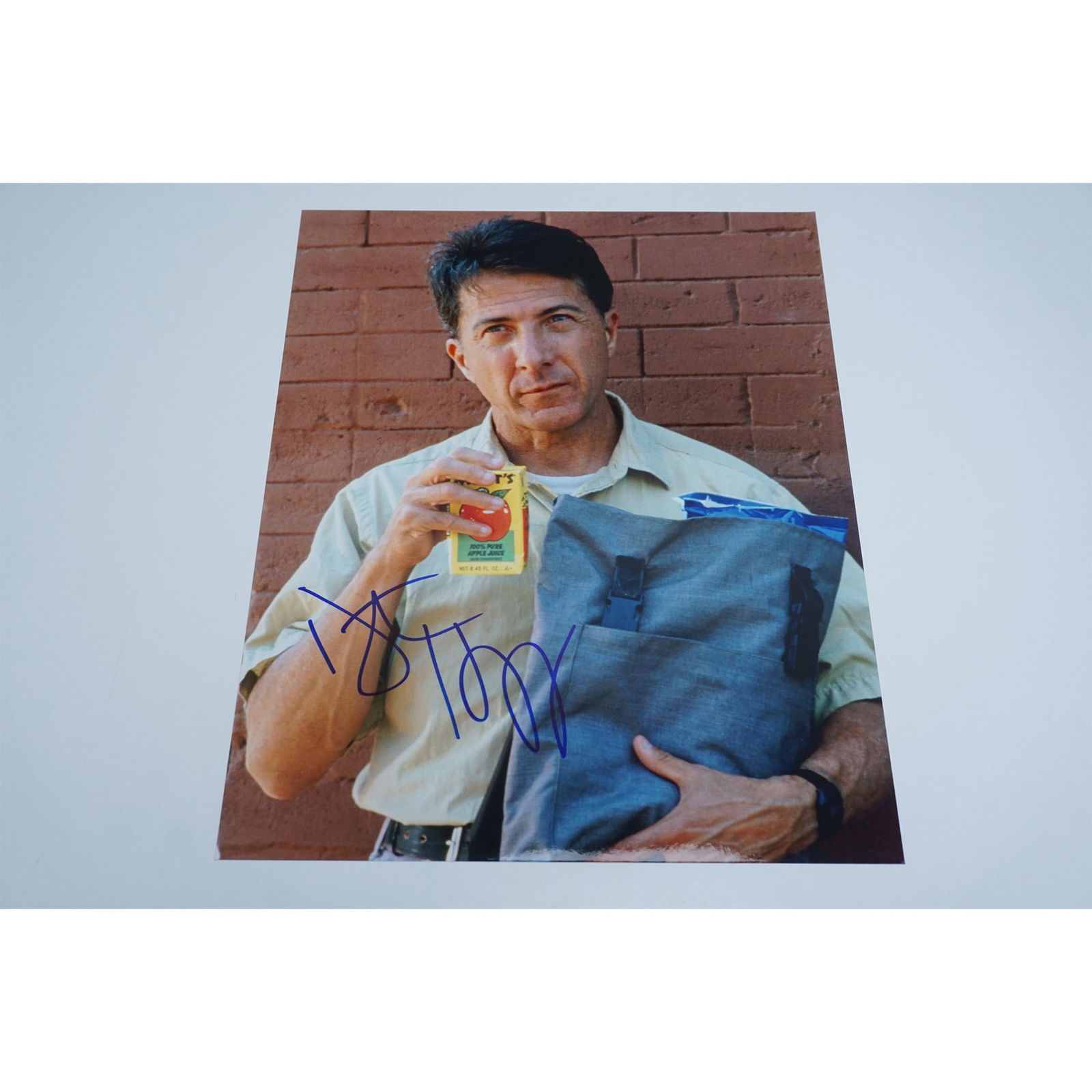 Dustin Hoffman Signed Photograph with COA: An 8x10 color photograph of acclaimed actor Dustin Hoffman, depicting him holding a blue cloth bag and a container of apple juice. Signed by Hoffman in bold blue ink, the autograph is prominently plac