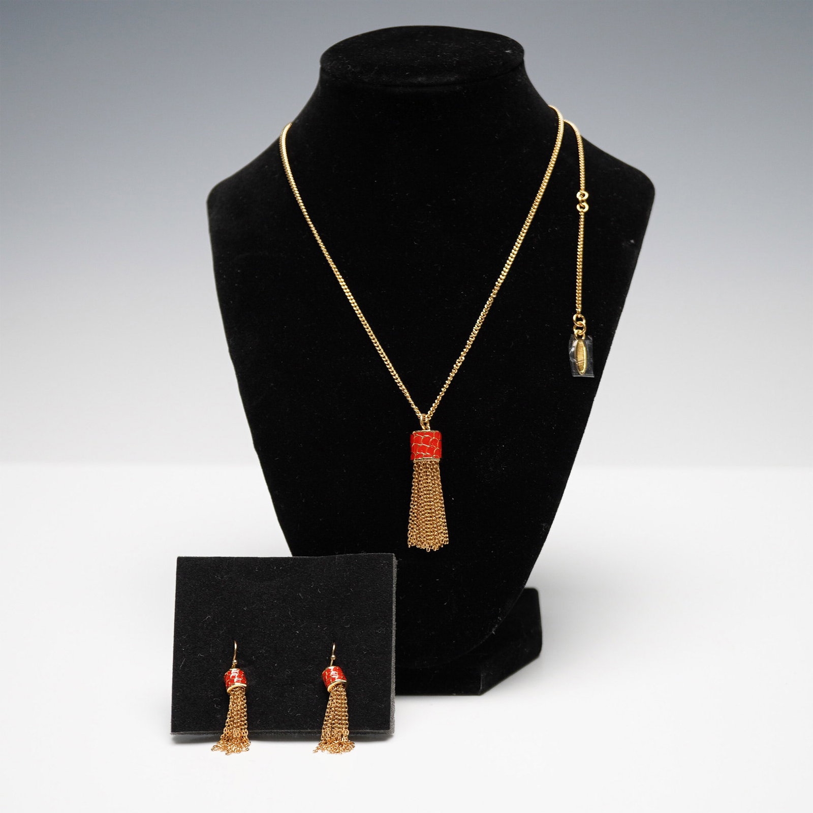 Halcyon Days Gold-Tone Tassel Necklace and Earring Set: Halcyon Days gold-tone necklace and earring set featuring a tassel design with intricate chain strands. The necklace showcases a red enamel-patterned cylindrical accent from which multiple gold-tone c