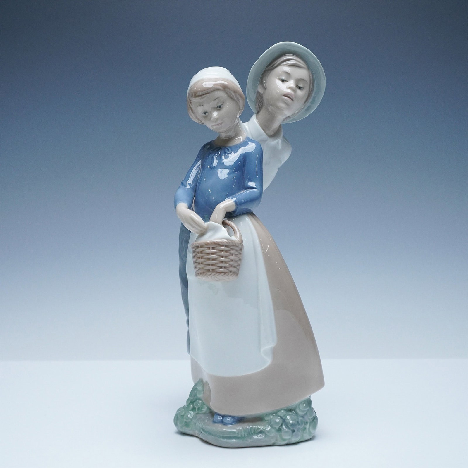 NAO by Lladro Porcelain Figurine, Goody Basket: Charming NAO by Lladro porcelain figurine titled Goody Basket. This beautifully sculpted piece captures a tender moment between a young boy and girl, with the boy leaning in affectionately as the girl