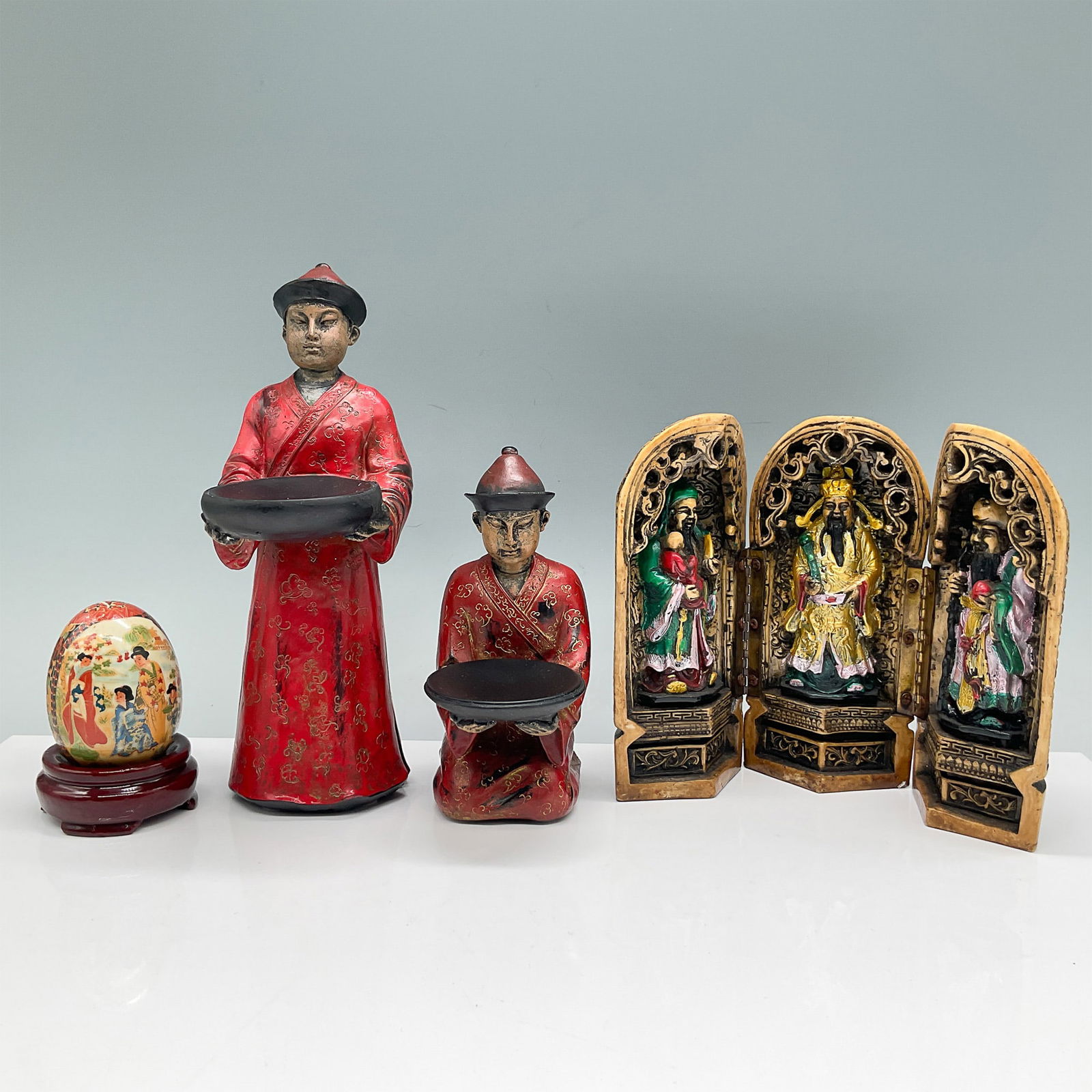 4pc Chinese Figurines and Decorative Egg with Stand: This lot includes four pieces of Chinese-inspired decorative art. The set features two hand-painted red-clad figures, one standing (8"H) and one seated, each holding a black bowl. Additionally, the lo