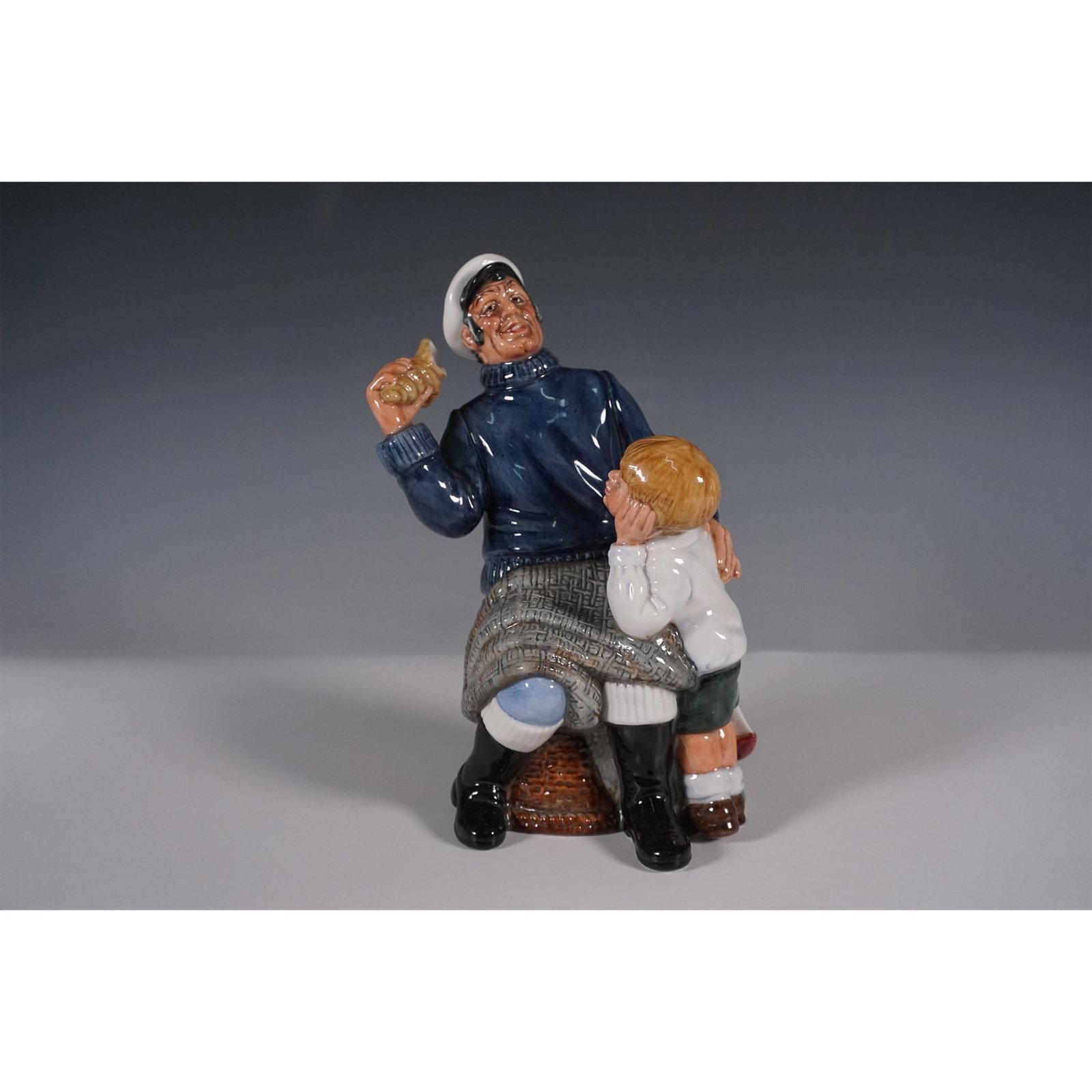Royal Doulton Figurine, Song of the Sea HN2729: This Royal Doulton figurine, titled Song of the Sea (HN2729), depicts a heartwarming maritime scene featuring a sailor seated on a wicker basket, holding a conch shell while sharing a moment with a yo