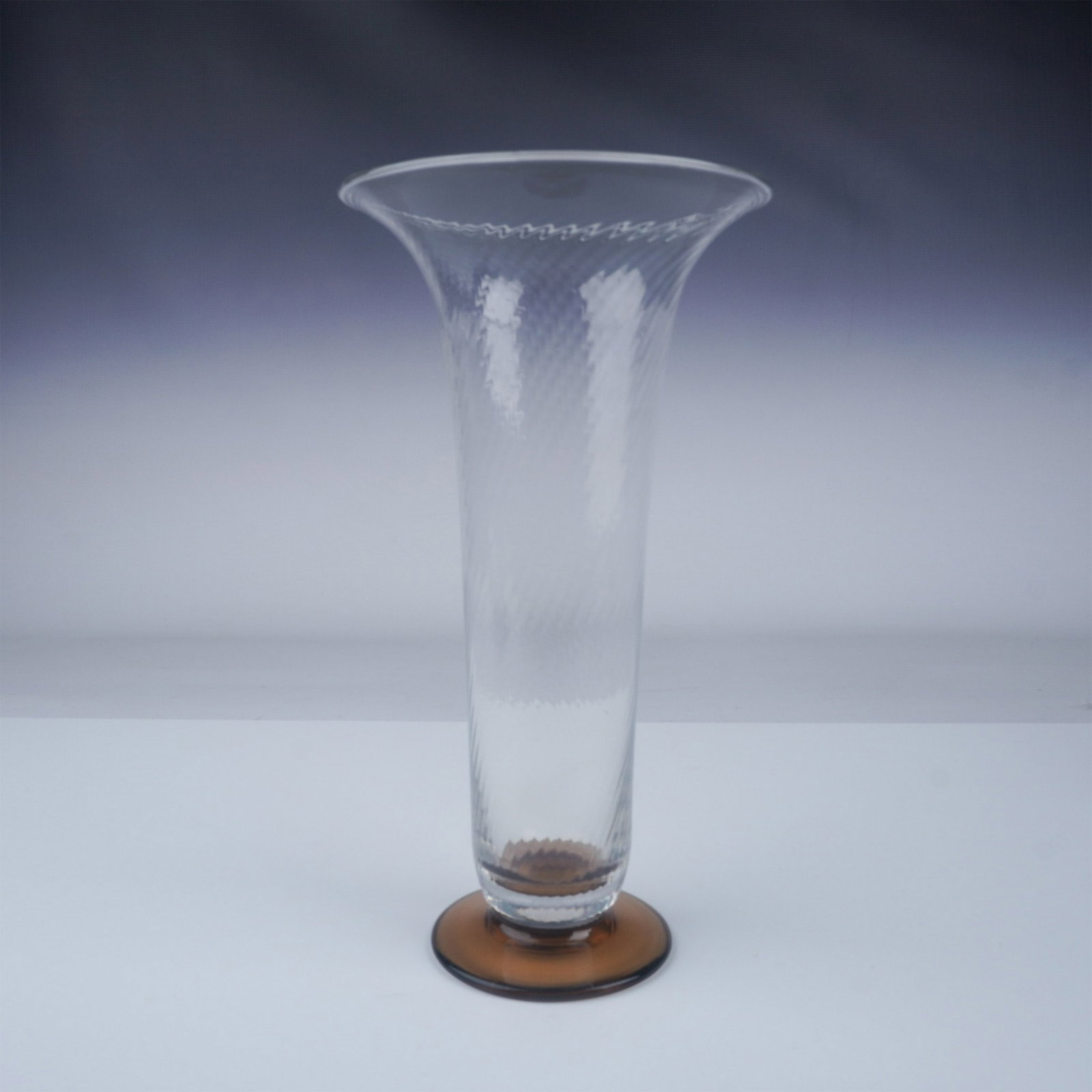 Vintage Glass Trumpet-Shaped Vase: A tall, glass vase featuring a flared rim and a subtle, embossed swirl pattern that adds texture and depth to the surface. The base is finished in a warm amber hue, providing a striking contrast to th