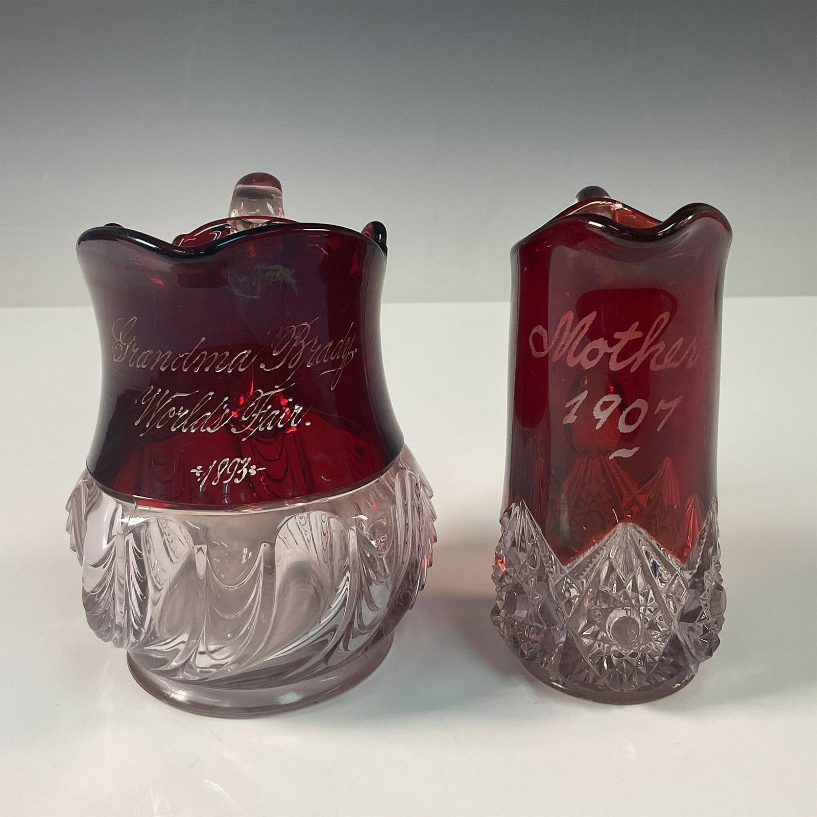 Pair of Vintage Red Glass Souvenir Creamers: A pair of antique red and clear glass souvenir creamers, each steeped in historical charm. The first creamer, inscribed "Grandma Brady World's Fair 1893," is a remarkable keepsake from the Chicago Wor