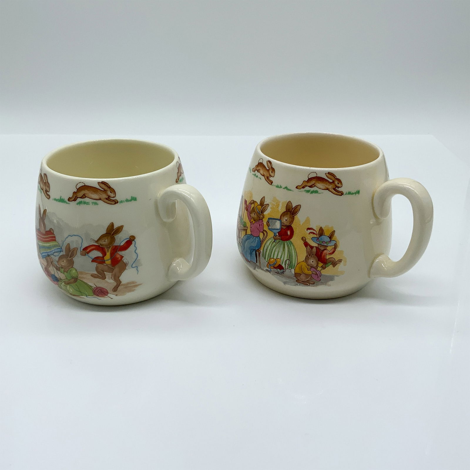 2pc Royal Doulton Bunnykins Mugs, Childrens Cups: his lot features a delightful pair of Royal Doulton Bunnykins mugs, each showcasing charming hand-painted scenes of playful rabbits in a variety of activities. These mugs, part of the Bunnykins series
