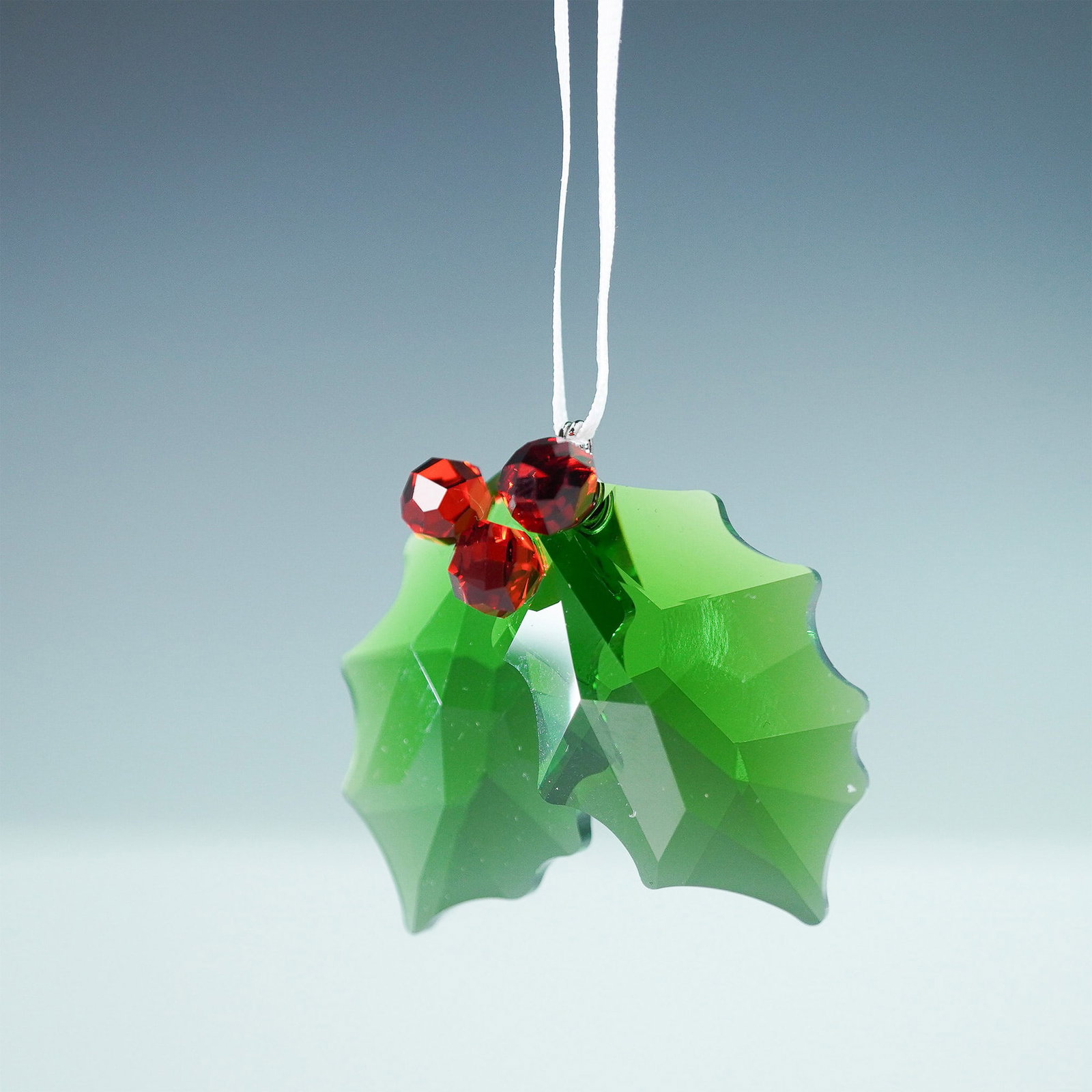 Swarovski Crystal Holly Ornament: This Swarovski crystal ornament features a holly design with green crystal leaves and red berry accents, suspended by a white ribbon. A festive addition to any holiday collection, it showcases Swarovs