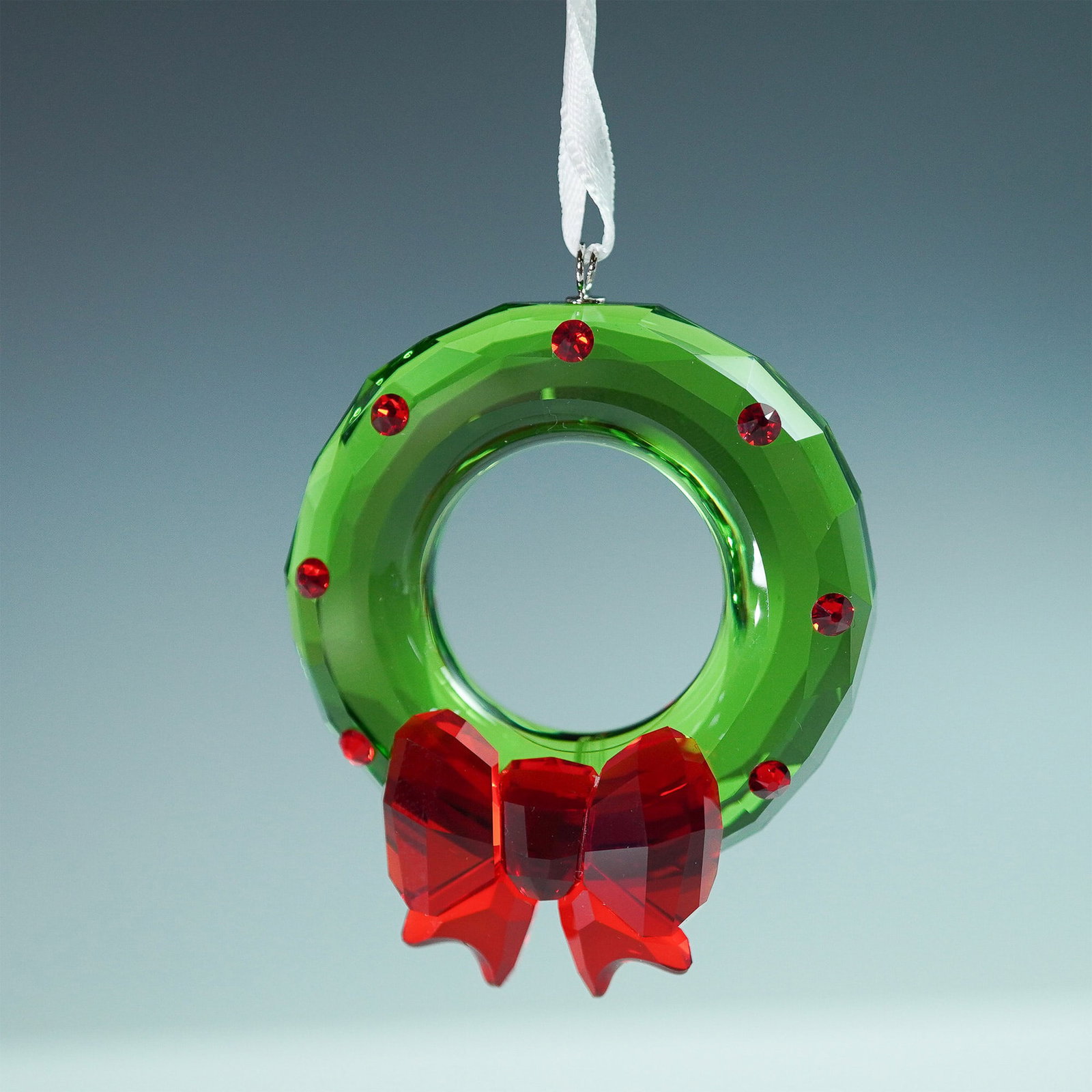 Swarovski Joyful Wreath Ornament: The Swarovski Joyful Wreath Ornament showcases a beautifully faceted green crystal wreath adorned with red crystal accents and a striking red crystal bow. Suspended from a white ribbon, this elegant h