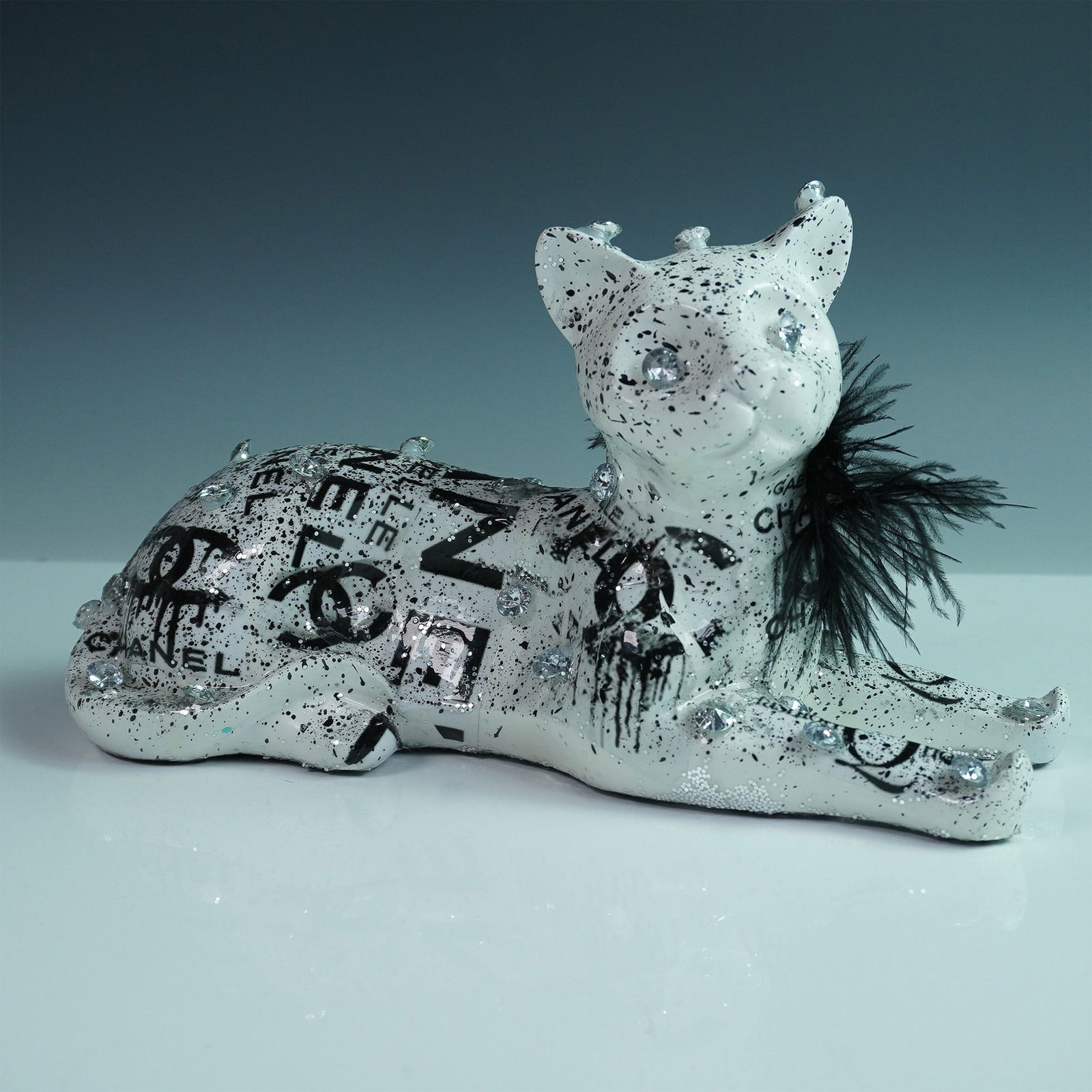 Khalil Douisse Mixed Media Cat Sculpture: A striking mixed media sculpture by contemporary artist Khalil Douisse, depicting a stylized cat in a reclining pose. The piece is adorned with black and white Chanel-inspired graffiti motifs, bold dr