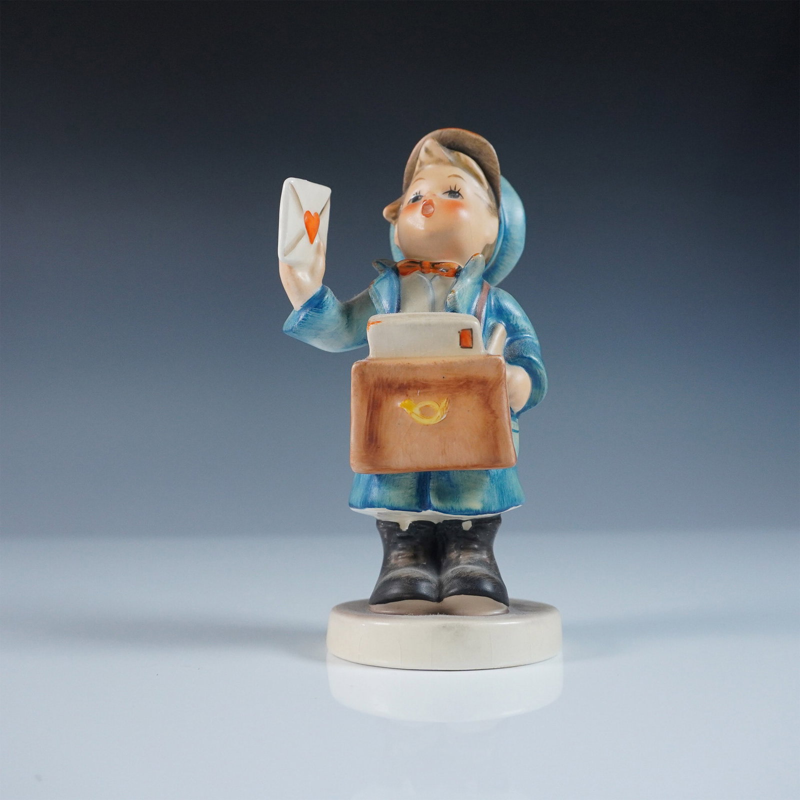 Goebel Hummel Figurine ? Mailman: Goebel Hummel figurine titled Mailman, featuring a young postal carrier dressed in a blue coat and hat, holding a letter with a red heart. With a satchel marked by a postal horn this charming piece em