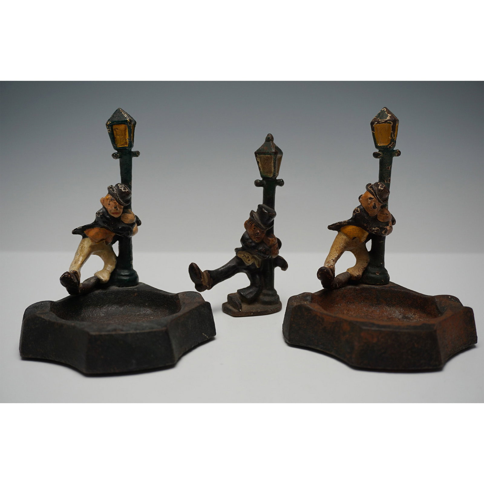 3pc Antique Cast Iron Drunkard Ashtrays and Match Holder Set: A set of three antique cast iron figural pieces featuring a humorous "drunkard" theme. Two of the pieces serve as ashtrays, with a drunken man slumped against a street lamp, while the smaller piece fu