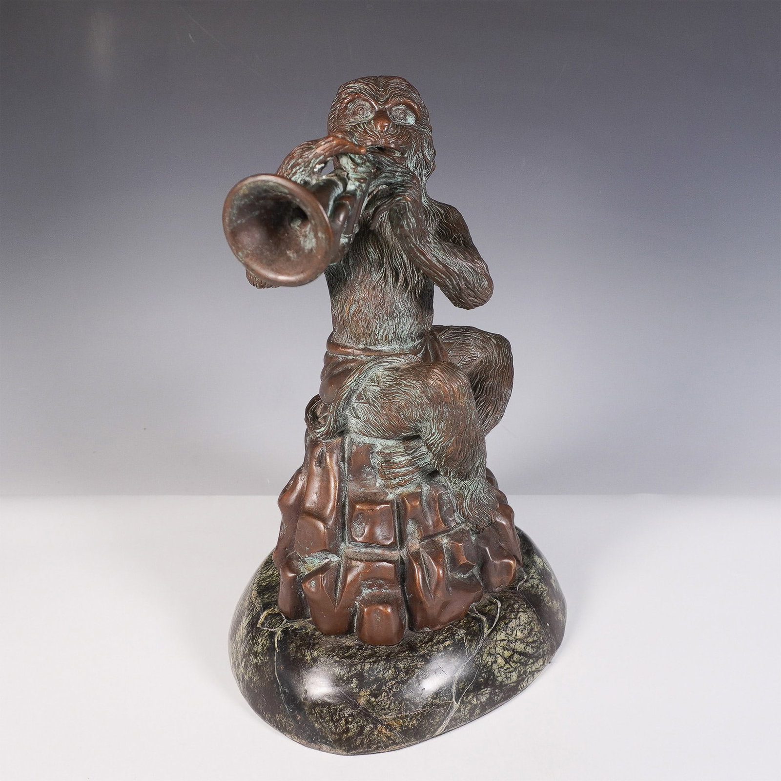 Theodore and Alexander Bronze Monkey Musician Sculpture: A finely crafted Theodore Alexander bronze sculpture depicting a monkey playing a trumpet. The figure is seated on a rock-like base, with intricate detailing in the fur texture and drapery. The sculpt