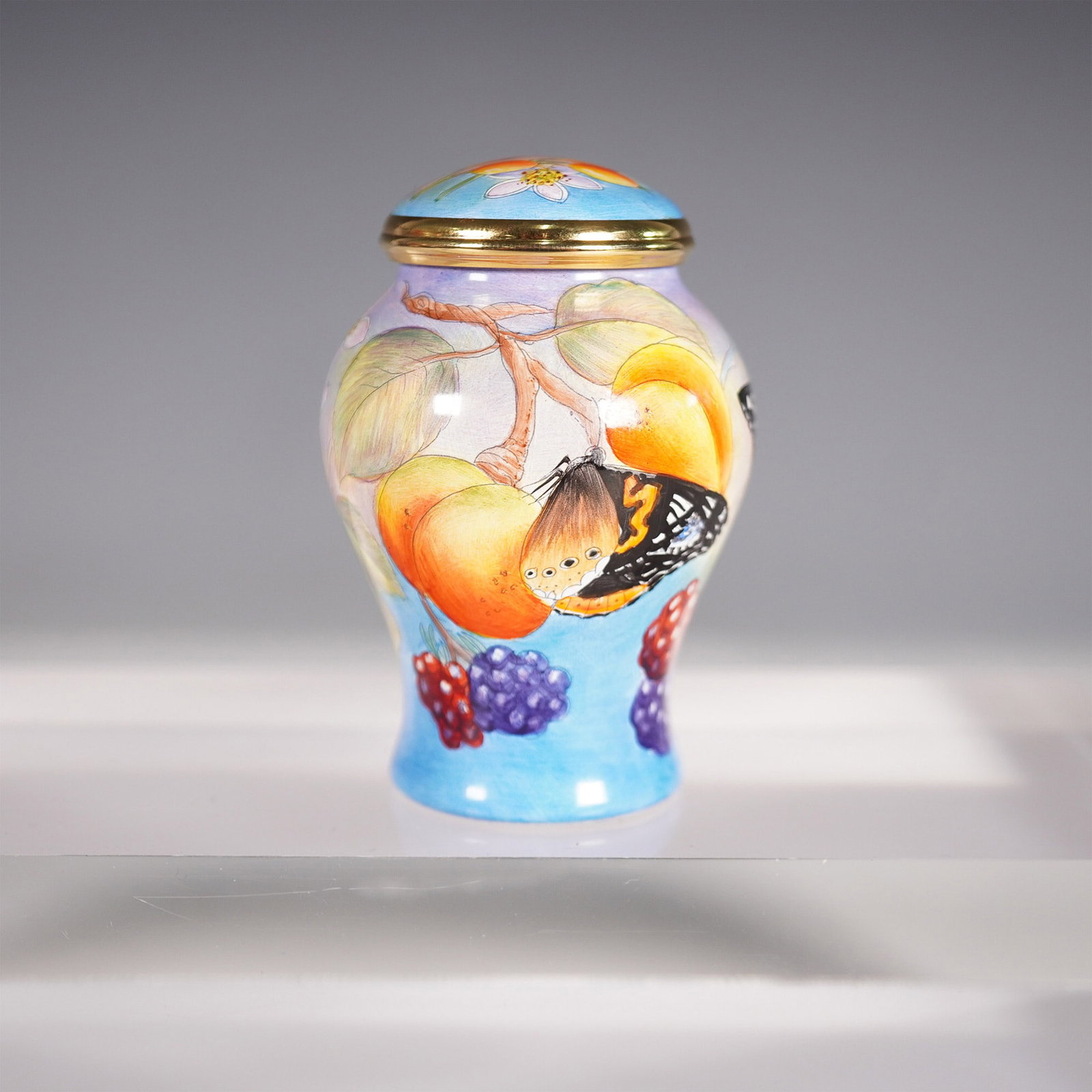 Moorcroft Small Enamel Ginger Jar, Butterfly & Fruit Design: A Moorcroft small enamel ginger jar featuring a vibrant hand-painted design of butterflies fluttering among ripened fruit and lush foliage. Set against a soft blue and lavender background, the intrica