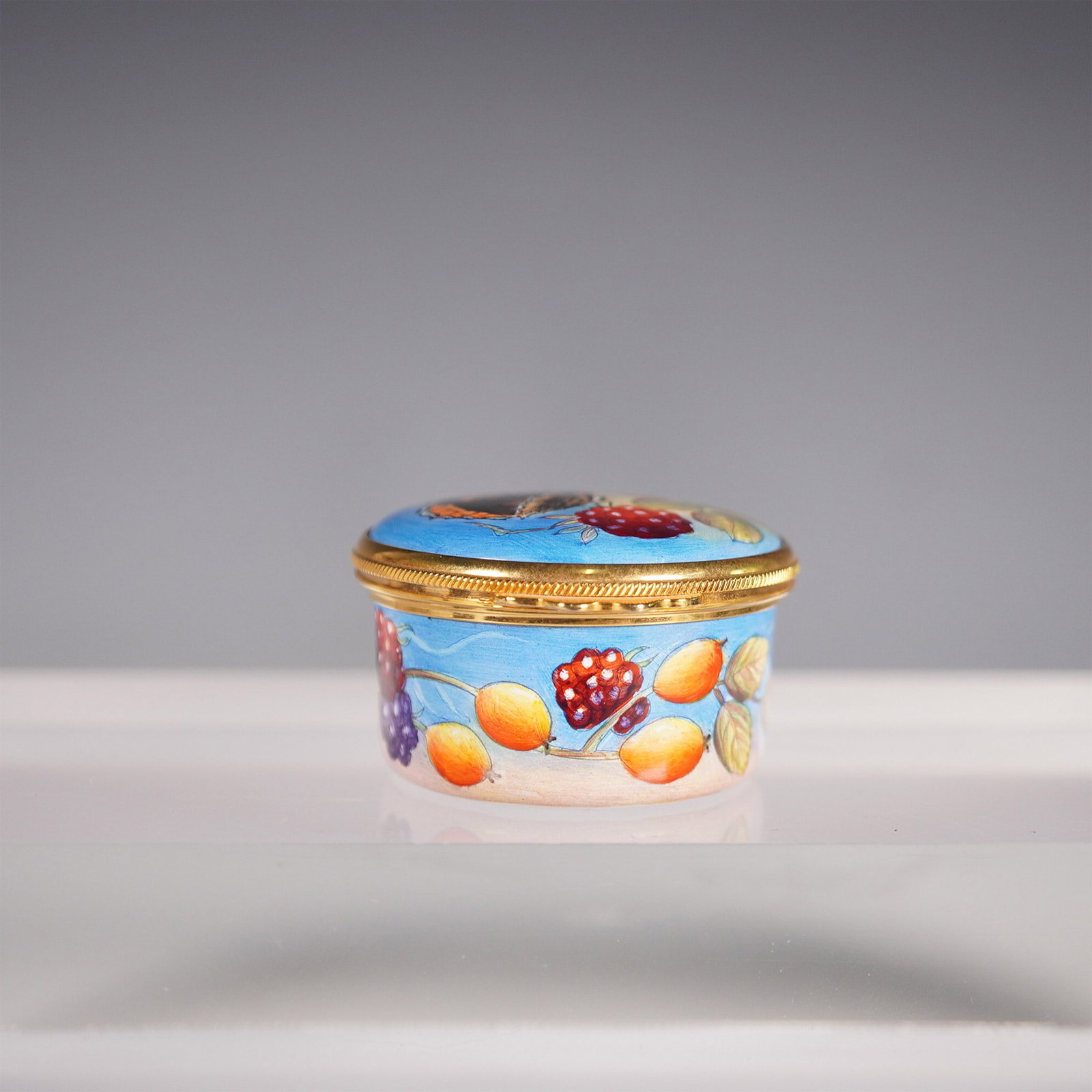 Moorcroft Small Oval Enamel Pot, Butterfly & Berry Design: A Moorcroft small oval enamel pot featuring a charming hand-painted design of ripe berries and fruit set against a soft blue background. The hinged lid showcases a beautifully detailed butterfly, whic