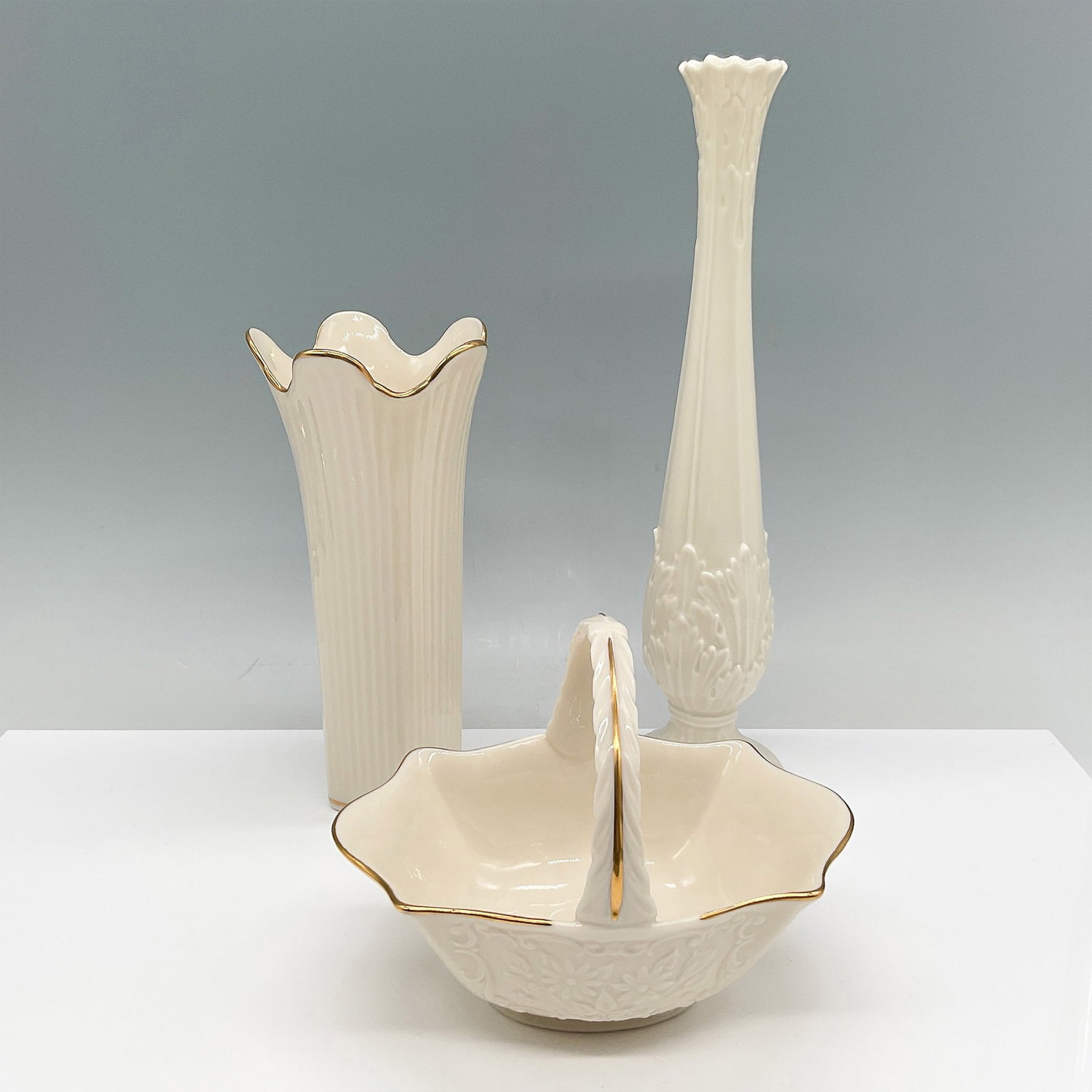 3pc Lenox Ivory Porcelain Vase and Basket Collection: A collection of three Lenox ivory porcelain pieces featuring elegant gold trim. The set includes a tall bud vase (11"H) with intricate embossed floral detailing, a fluted vase (7.25"H) with ribbed tex