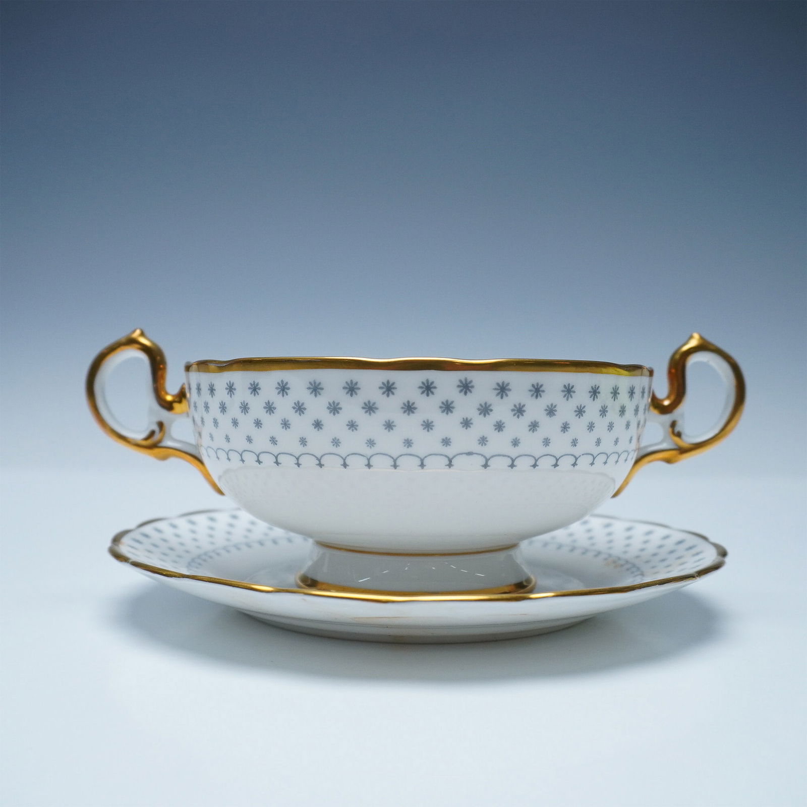 2pc Royal Chelsea English Bone China Gray & Gold Soup Cup and Saucer: A refined Royal Chelsea English bone china cream soup cup and saucer set, featuring an intricate blue starburst pattern accented with rich gold trim. The elegantly shaped cup showcases ornate double h