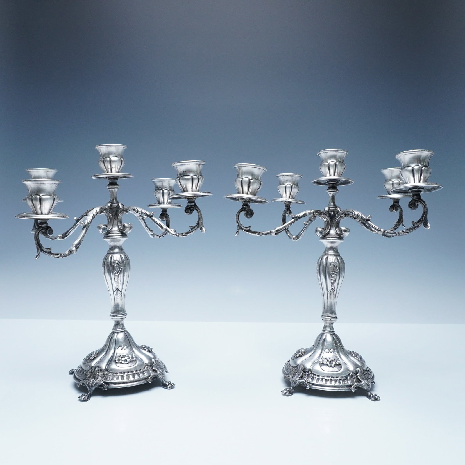 Pair of Italian Handmade Ornate Silver-Plated Candelabras: Elegant pair of Italian silver-plated candelabras, each featuring five candle holders with gracefully curved arms and intricate detailing. The bases are adorned with embossed floral and scroll motifs,