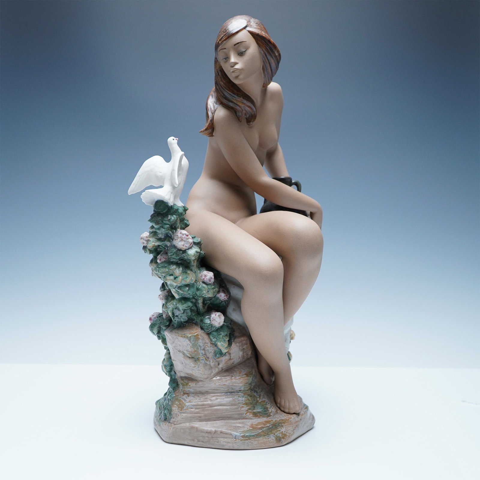 Lladro Gres Nude with Dove Sculpture 01013503: A captivating Lladro porcelain figurine titled Nude with Dove, model number 01013503. This elegant sculpture features a seated nude woman with flowing hair, gazing at a white dove perched on a floral-