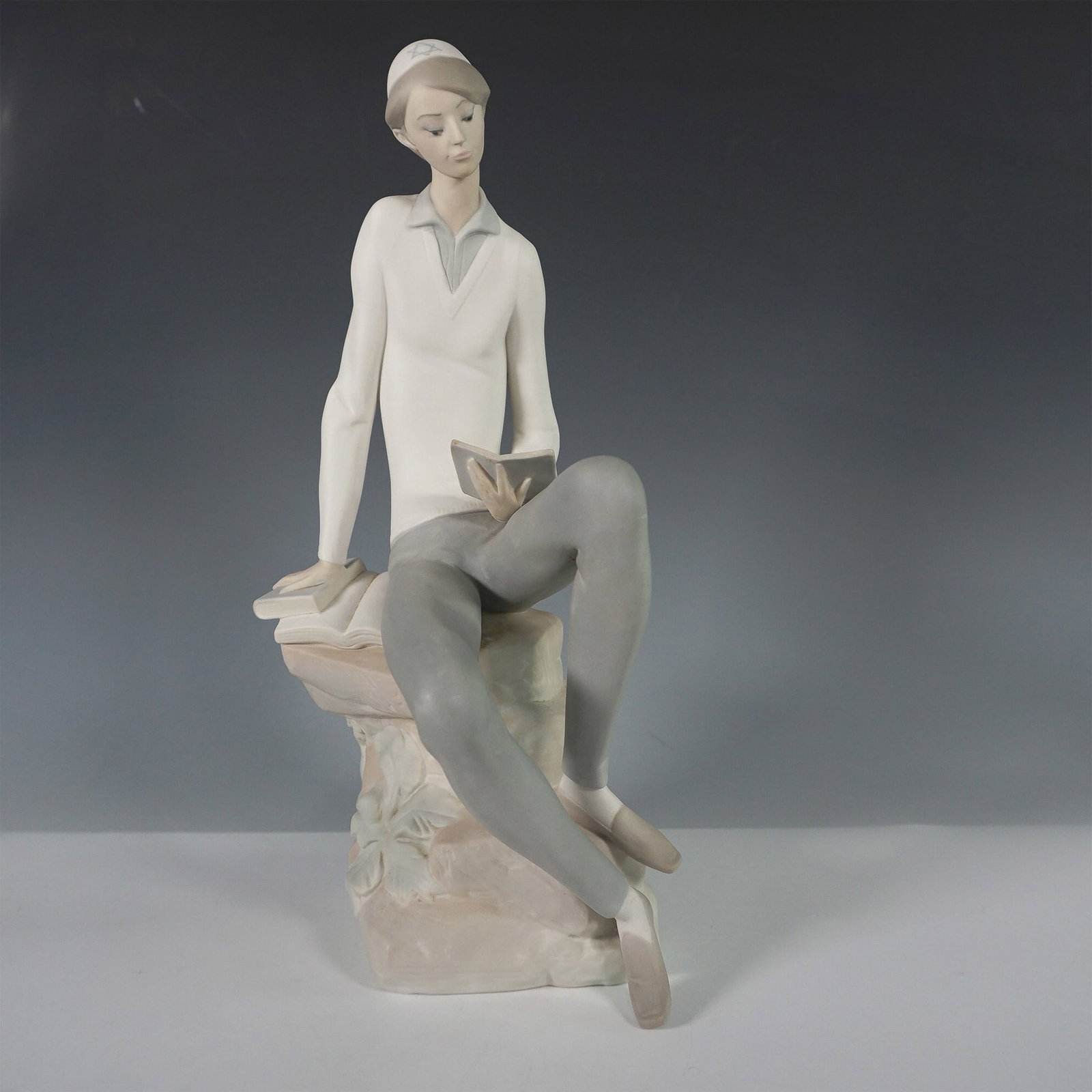 Lladro Porcelain Figurine, Hebrew Student 1014684: Exquisite Lladro porcelain figurine titled Hebrew Student, model number 1014684. This finely crafted piece depicts a young scholar seated on a stone, engrossed in reading a book. The figurine showcase