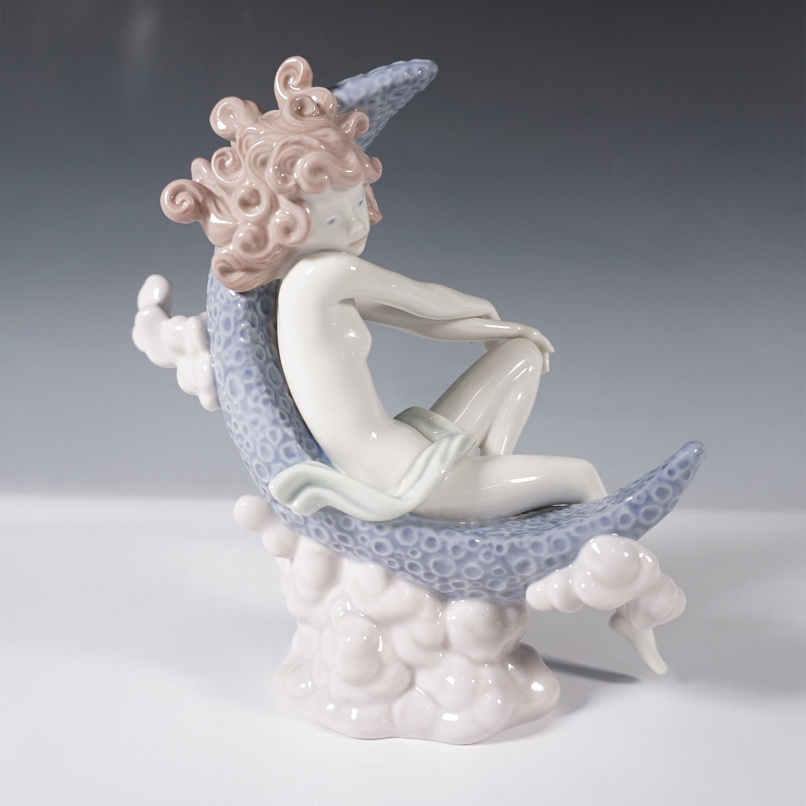 Lladro Porcelain Figurine, Moonlight 1001437: This figure features a delicate female figure seated within a crescent moon, surrounded by soft cloud-like details. Her flowing curls and serene pose evoke an ethereal presence. Finished in Lladro's s