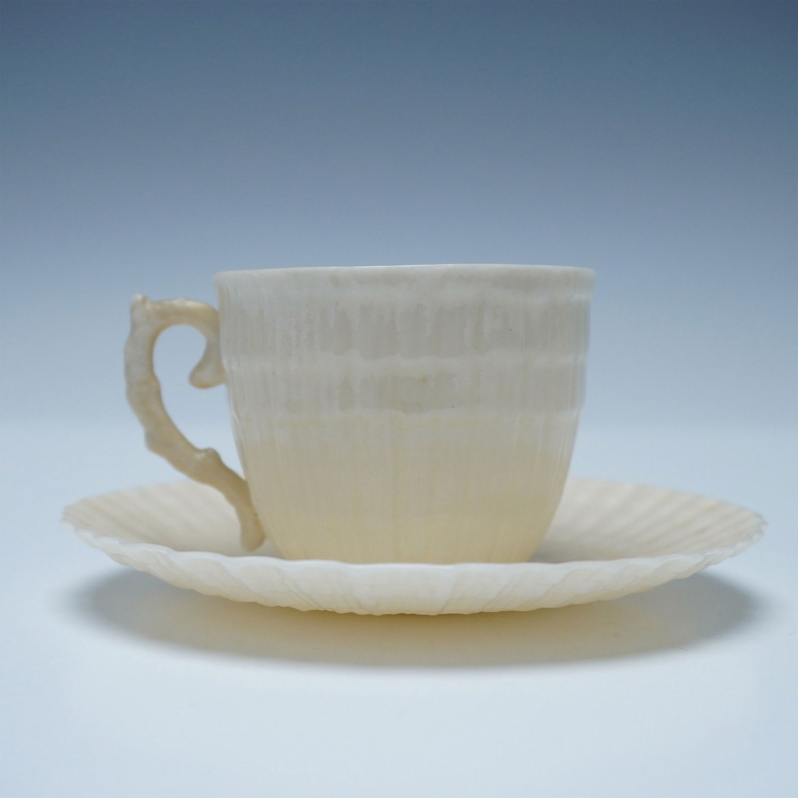 2pc Belleek Demitasse Cup and Saucer Set, Limpet: Two-piece set featuring a cream-colored espresso cup and saucer. The design includes a textured, embossed rim. Marked with Belleek backstamps and Reg No. 0857. Cup: 2"H. Saucer: 4.25"W x 0.5"H. Issued