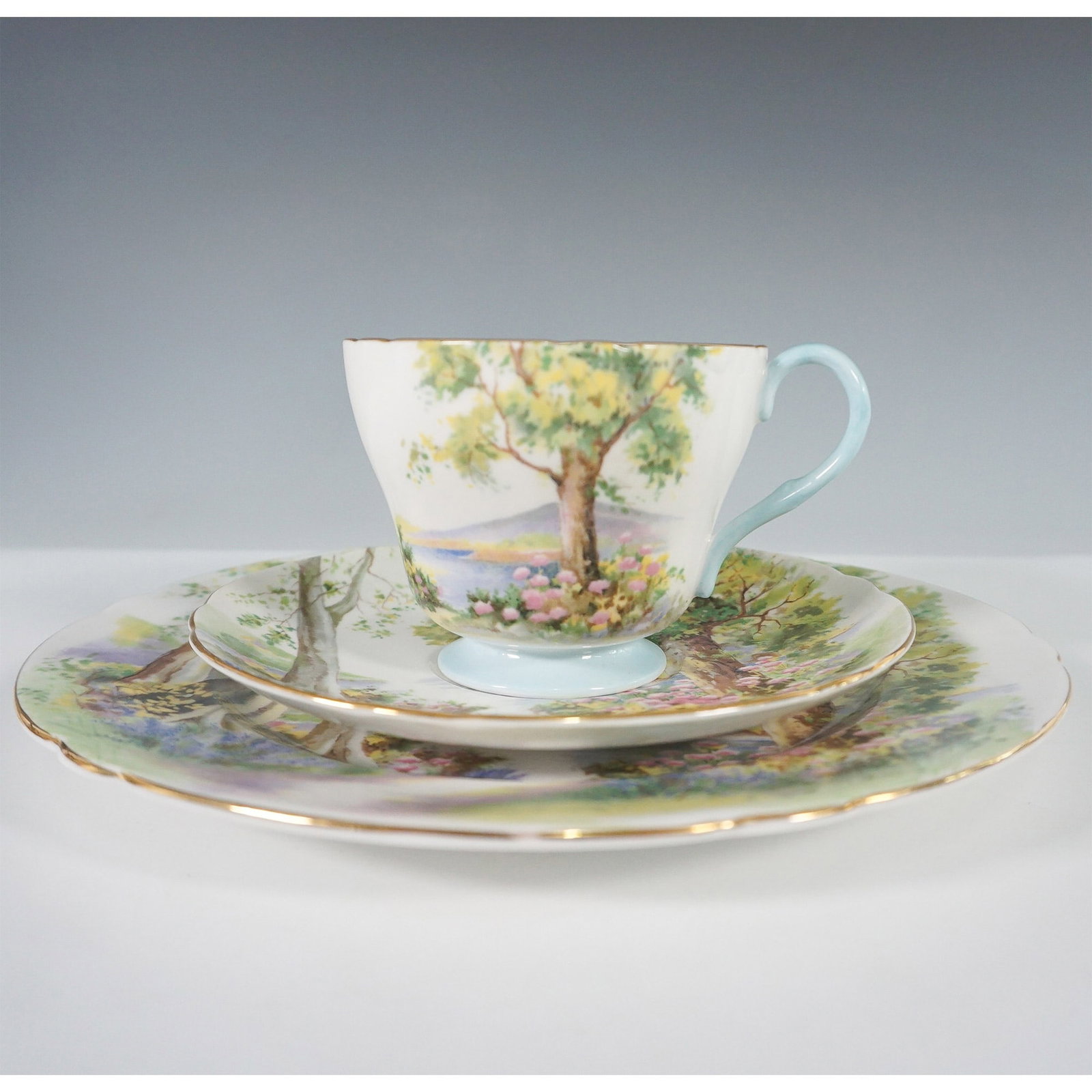 3pc Shelley England Woodland Trio ? Cup, Saucer, and Plate: Shelley England Woodland trio set, featuring a teacup, saucer, and plate adorned with a picturesque forest scene. The design captures a tranquil wooded landscape with towering trees, blooming flowers,