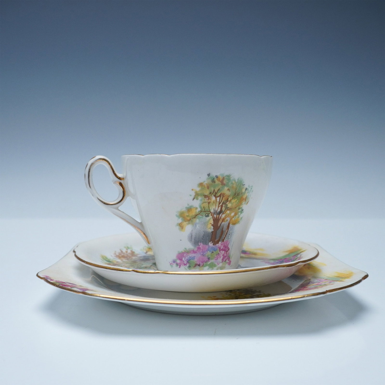 3pc Shelley Trio Tea Set, Englands Charm: A three-piece Shelley fine bone china tea set from the Ideal Series, featuring a hillside landscape design. The set includes a teacup, saucer, and tea plate, each with a scenic depiction of a tree-lin