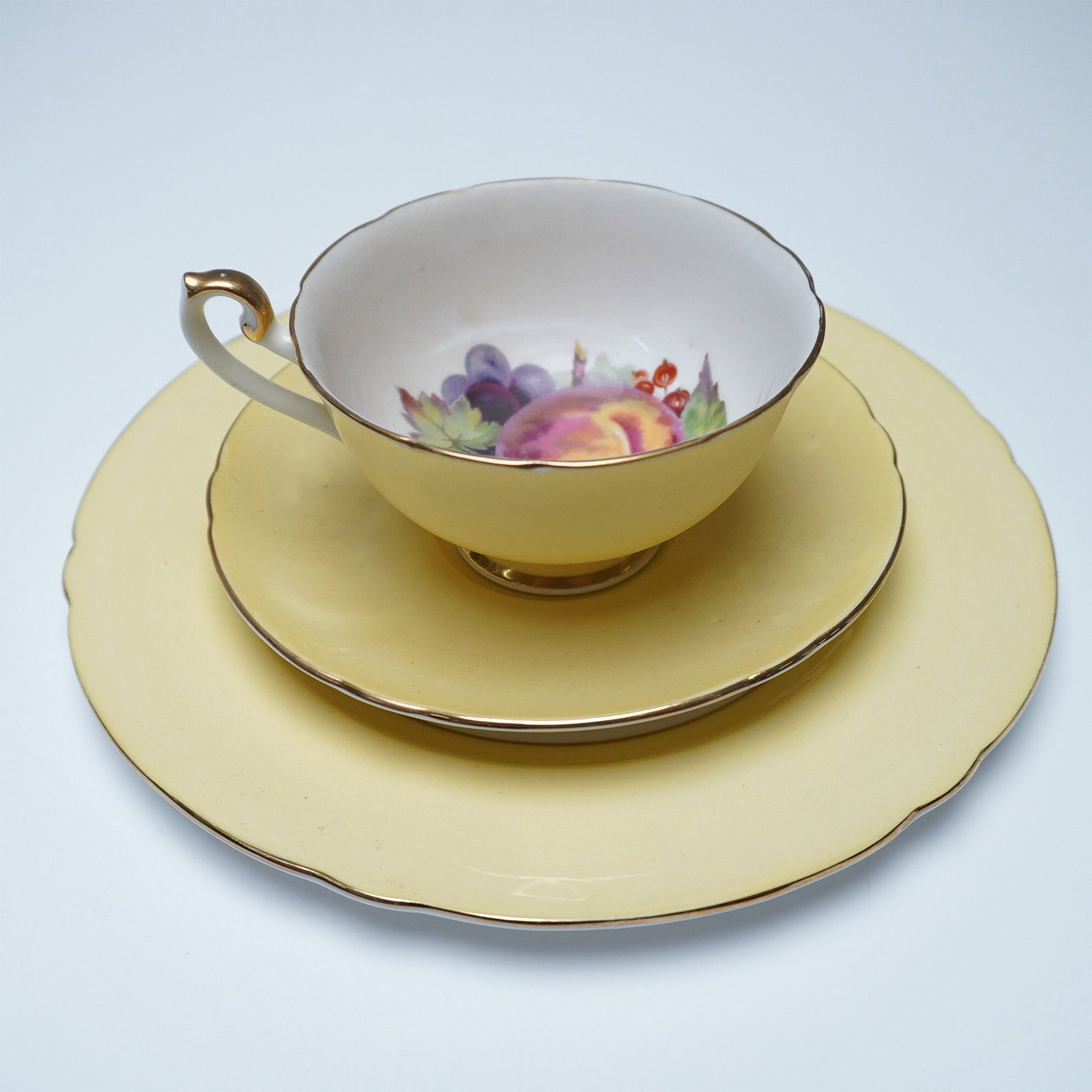 3pc Shelley Yellow Fruit Motif Trio Tea Set: A three-piece Shelley fine bone china tea set featuring a yellow exterior and a fruit motif on the interior. The set includes a teacup, saucer, and tea plate. Each piece with gilt trim. Marked with Sh