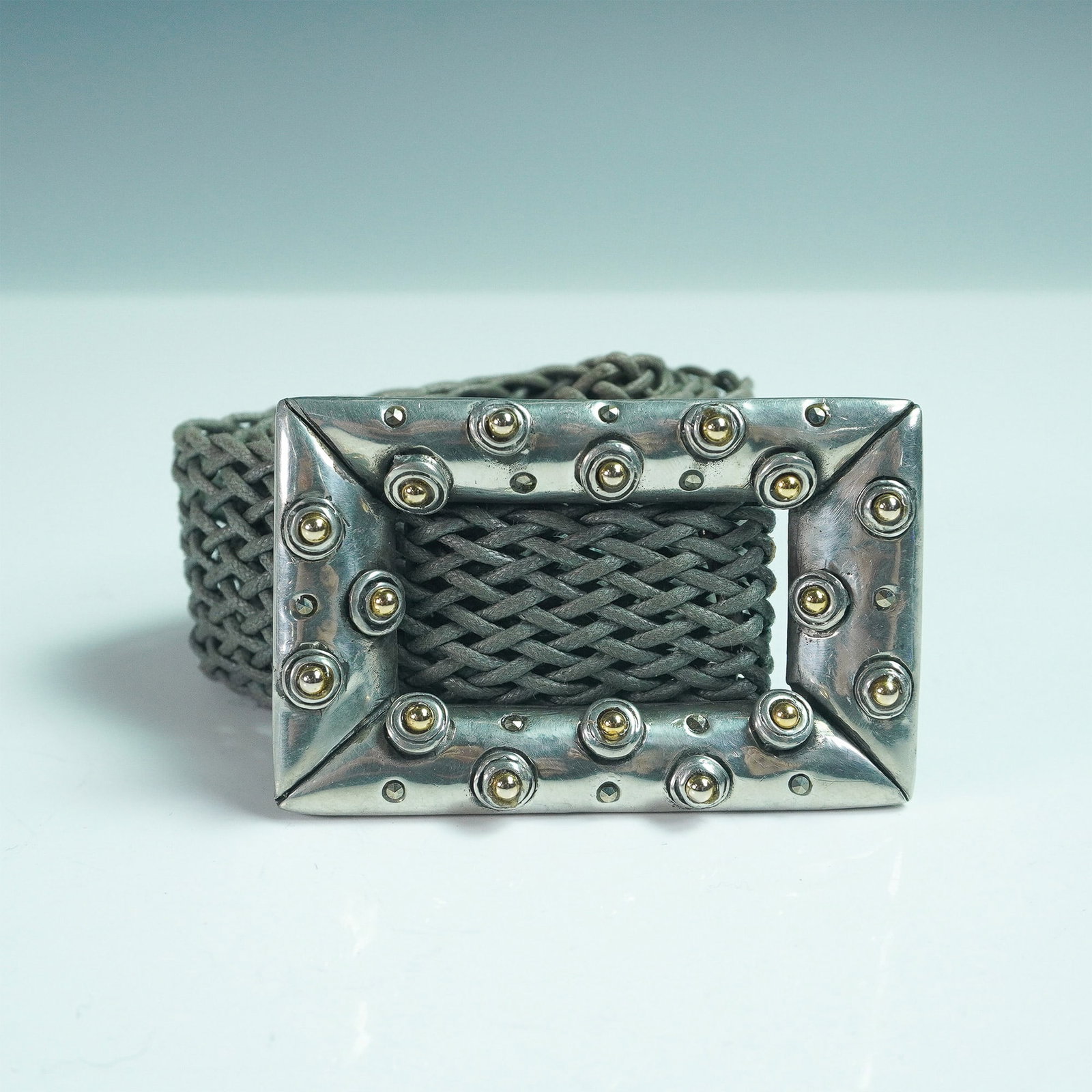 Connie Bates Fahrenheit Woven Cord Belt with Studded Metal Buckle: A striking Connie Bates Fahrenheit belt featuring a wide, intricately woven gray cord strap paired with a bold, studded metal buckle. The rectangular silver-tone buckle is accented with raised brass-t