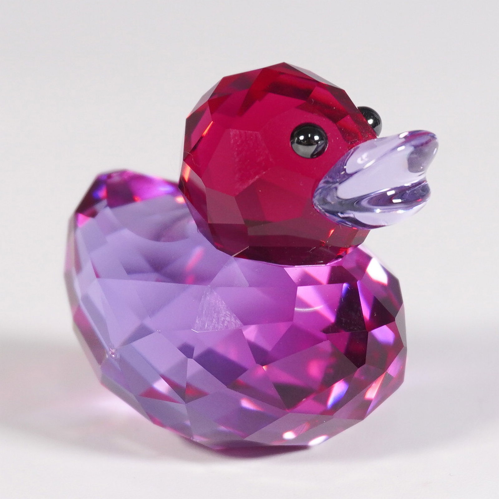 Swarovski Happy Ducks Crystal Figurine w/Original Box: A vibrant Swarovski crystal figurine from the Happy Ducks collection. This whimsical design features a rich pink and purple color gradient with a faceted body that sparkles under light. The duck has a
