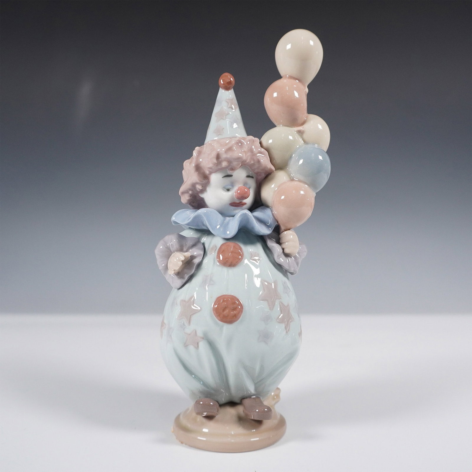 Lladro Porcelain Figurine, Littlest Clown 1005811: Charming Lladro porcelain figurine titled Littlest Clown (5811), sculpted by Antonio Ramos. This delightful piece depicts a young clown in pastel-colored attire, holding a vibrant cluster of balloons.