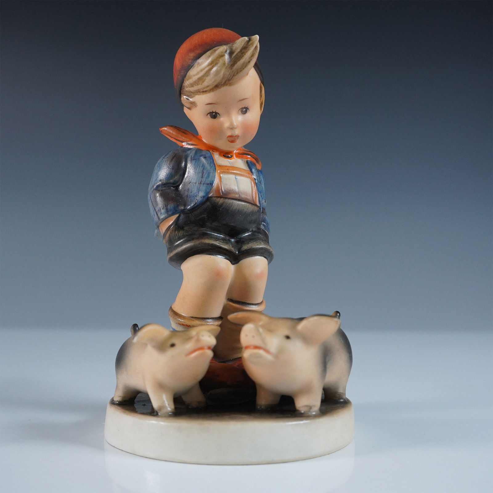 This Goebel Hummel Porcelain Figurine, Farm Boy: This Goebel Hummel figurine, captures a young boy dressed in a blue jacket, black shorts, knee-high socks, and a cape standing alongside two piglets. Maked with the Goebel Hummel backstamp.66. Issued: