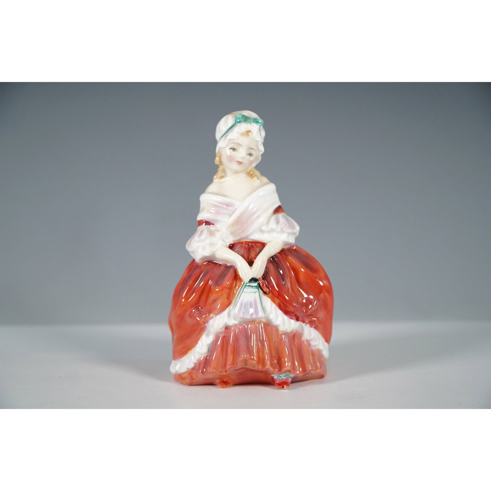Royal Doulton Bone China Figurine, Peggy HN2038: From the Harradine Child Classics series, this figurine depicts a young woman dressed in a striking red and white gown, gracefully holding a fan. Her delicate features, softly draped shawl, and bonnet